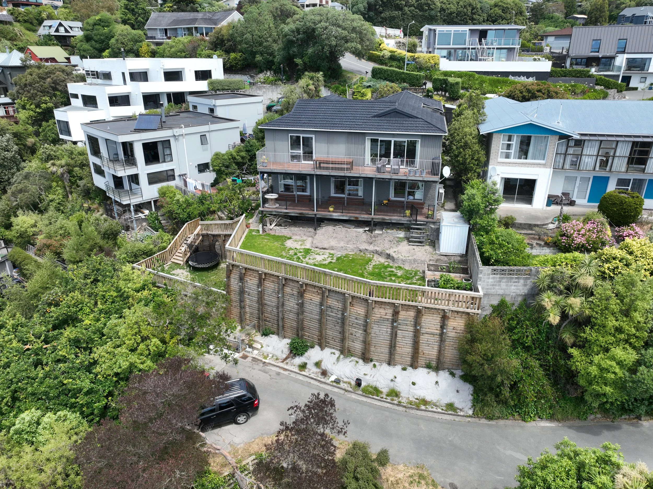 A neighborhood with multiple houses on a hillside, including a gray house with a balcony, a neighboring house with blue accents, and modern white houses, surrounded by trees and greenery, with a street and parked cars visible.