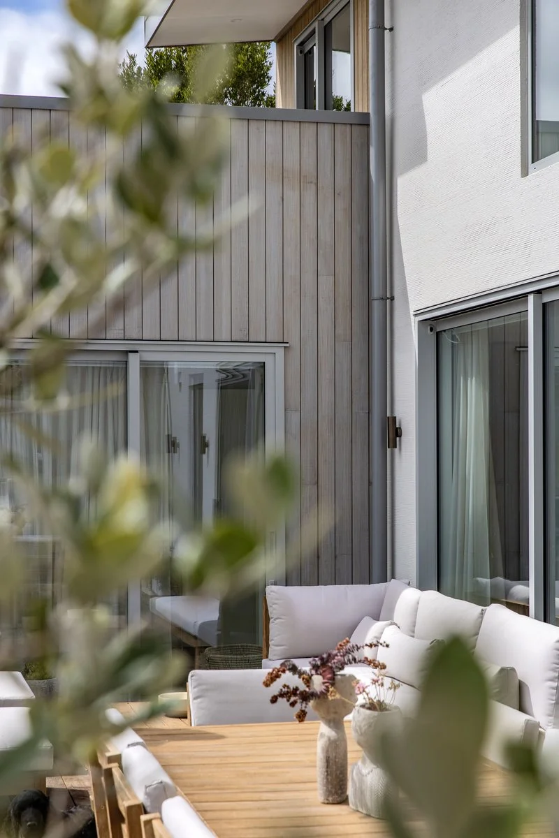 A backyard patio with a wooden table and white outdoor sofa, decorated with vases of dried and fresh flowers, surrounded by a modern house with large sliding glass doors and a wooden fence.
