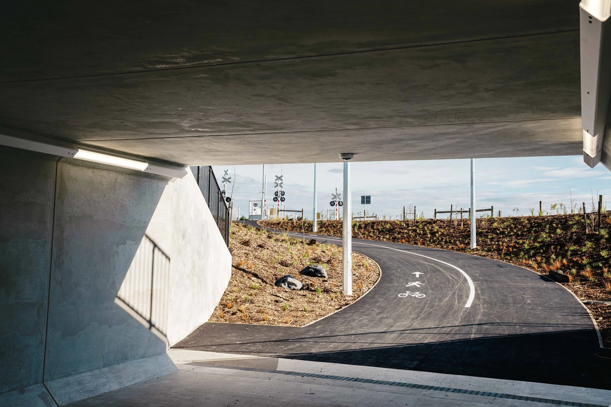 Underpass with a curved bike lane marked with a bicycle symbol, overlooking a rural scene with railroad crossing signs and light poles.