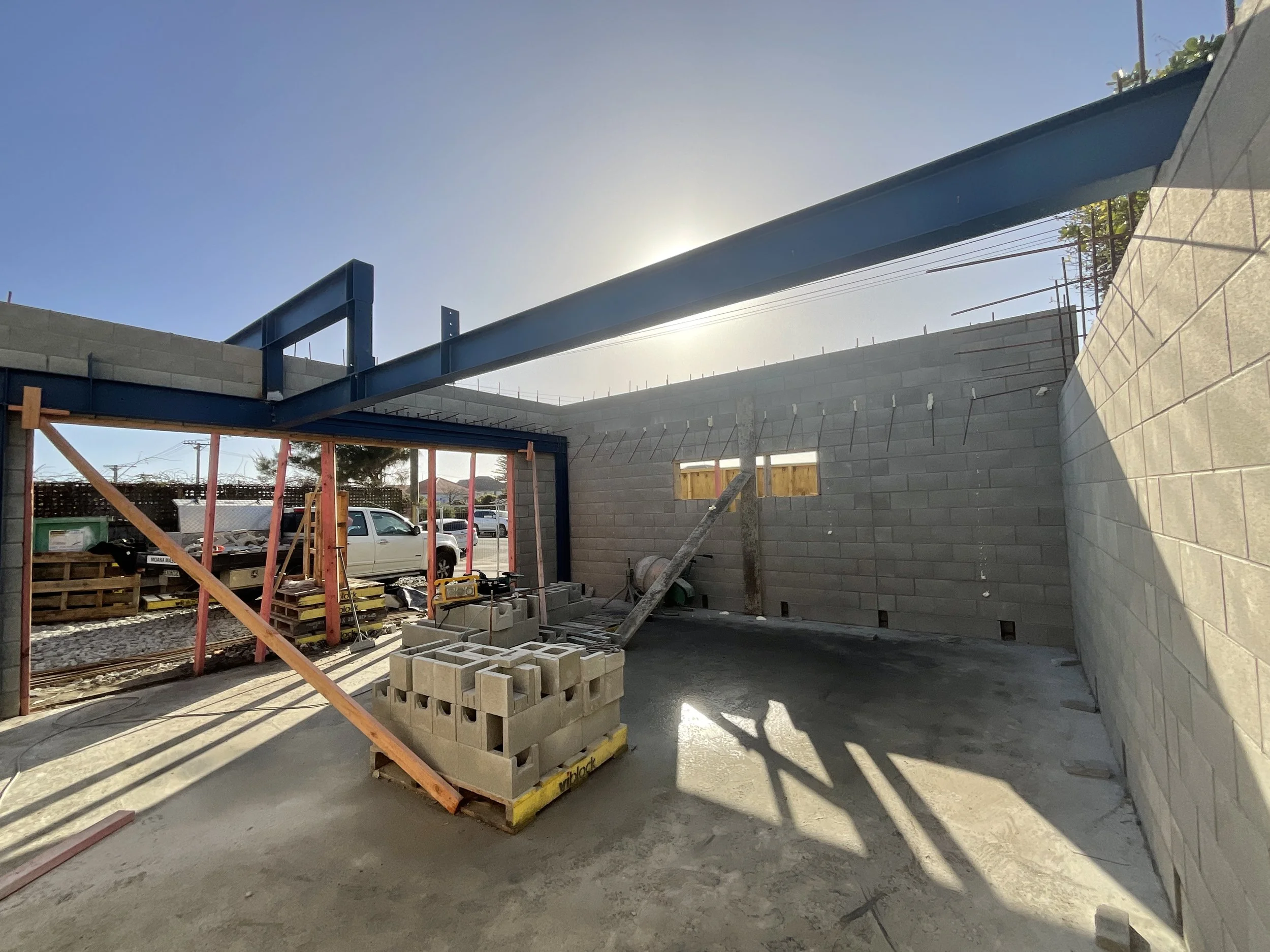 Construction site with unfinished concrete block walls, construction materials including bricks and scaffolding, and a large steel beam installed on top of the walls.