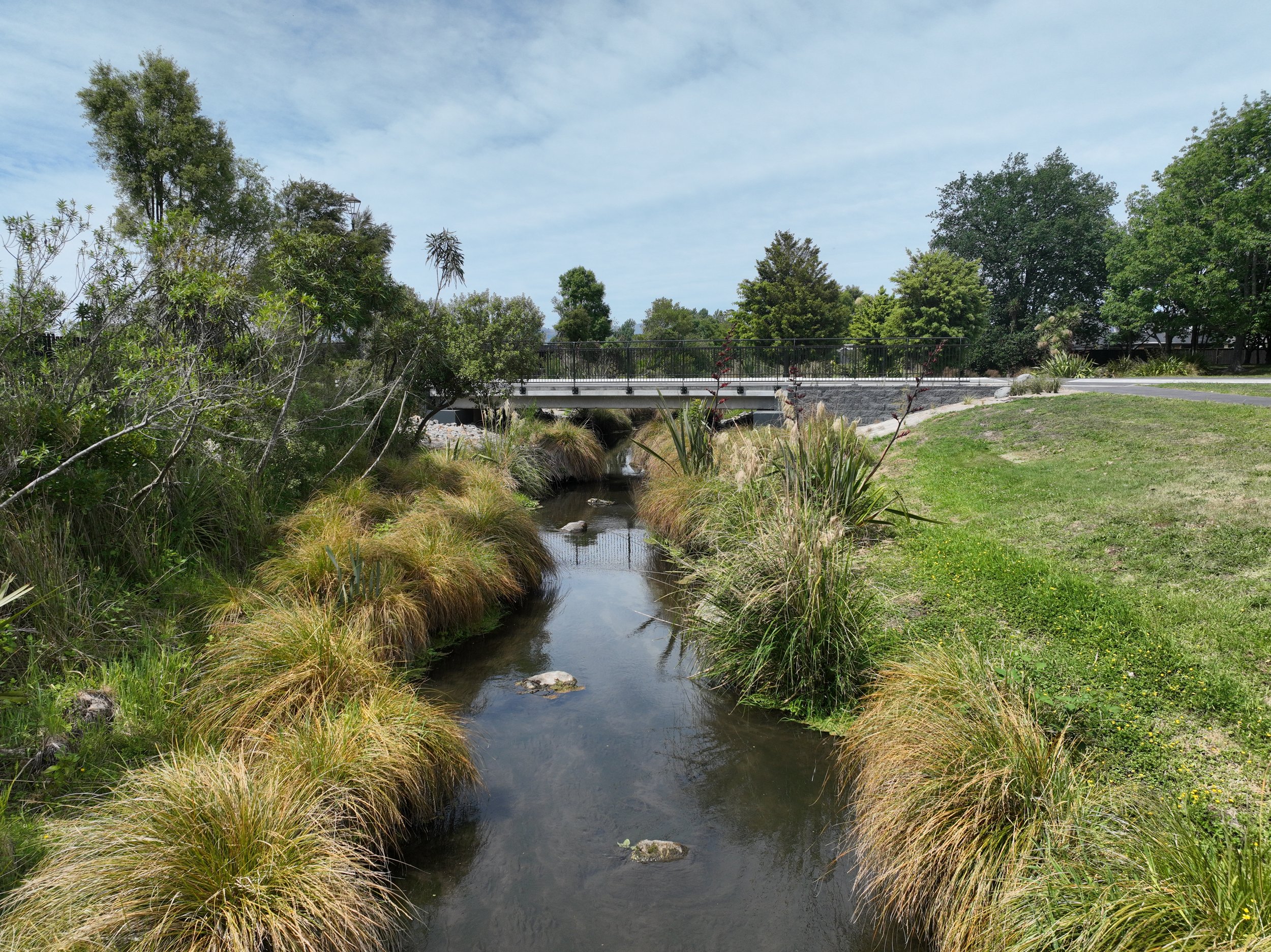 A small creek flowing under a bridge in a park with green trees and grassy areas on a partly cloudy day.