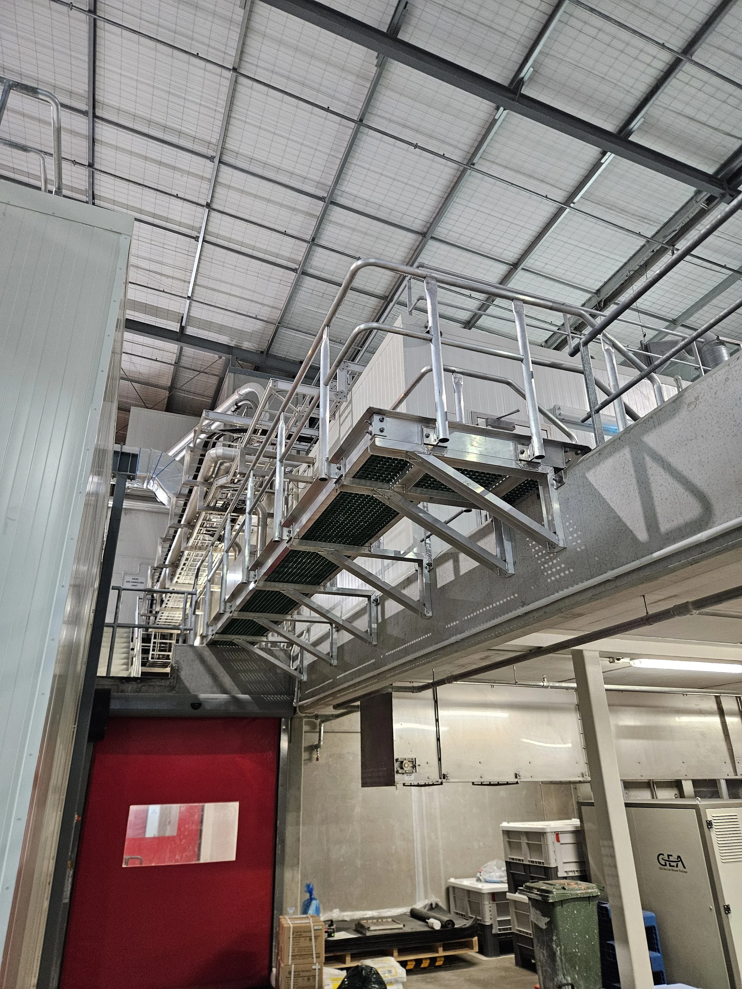 Interior of a warehouse or industrial facility with metal pipes, railings, and equipment. There are boxes, containers, and a red door.