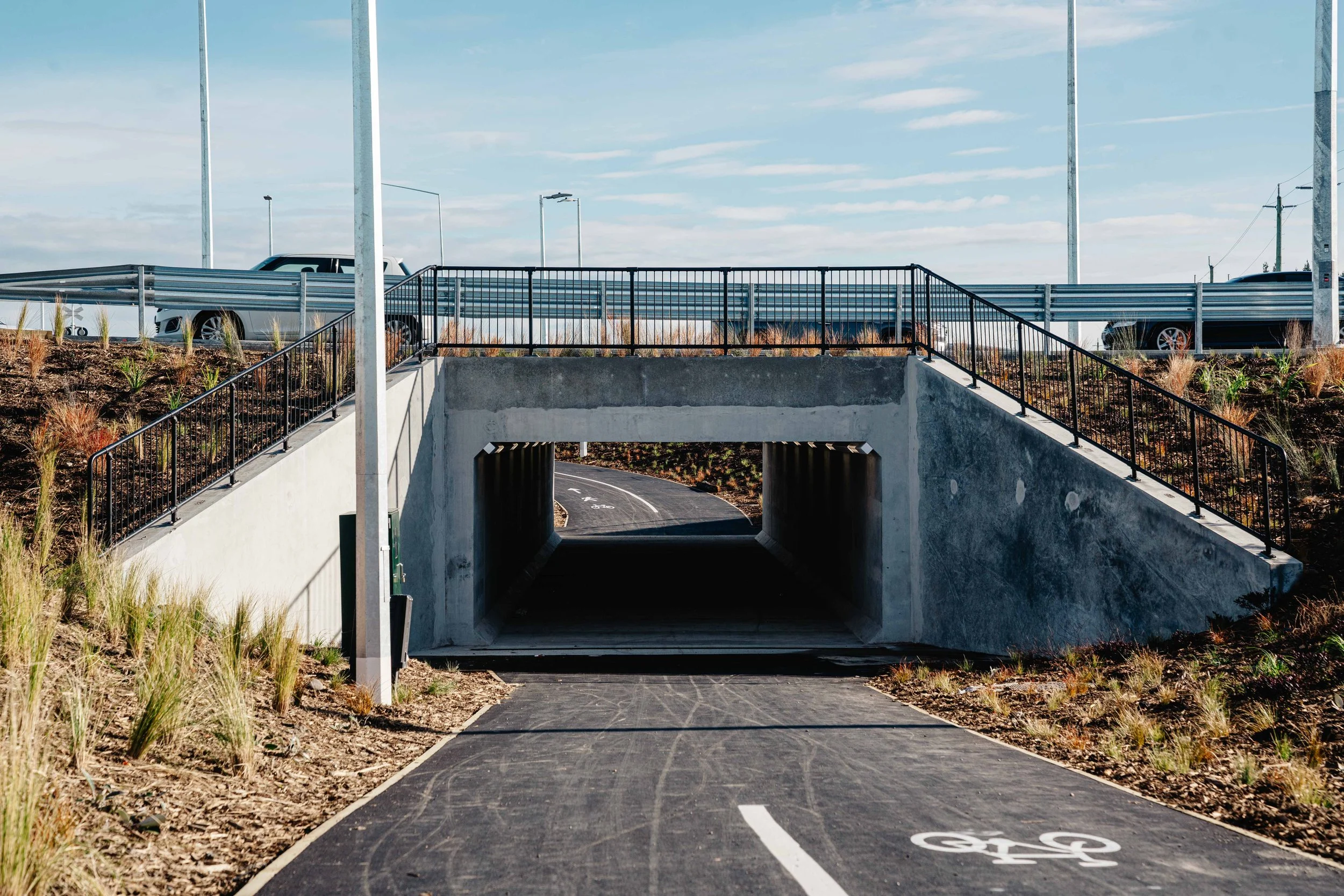 Underpass with a bike lane, fencing, and vehicles on the road above, with a blue sky and clouds.