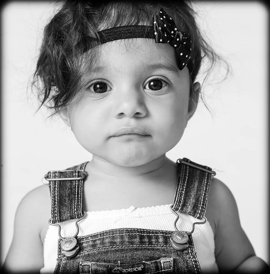 A black and white photo of a young girl with curly hair, wearing a headband with a bow, and denim overalls over a white top.