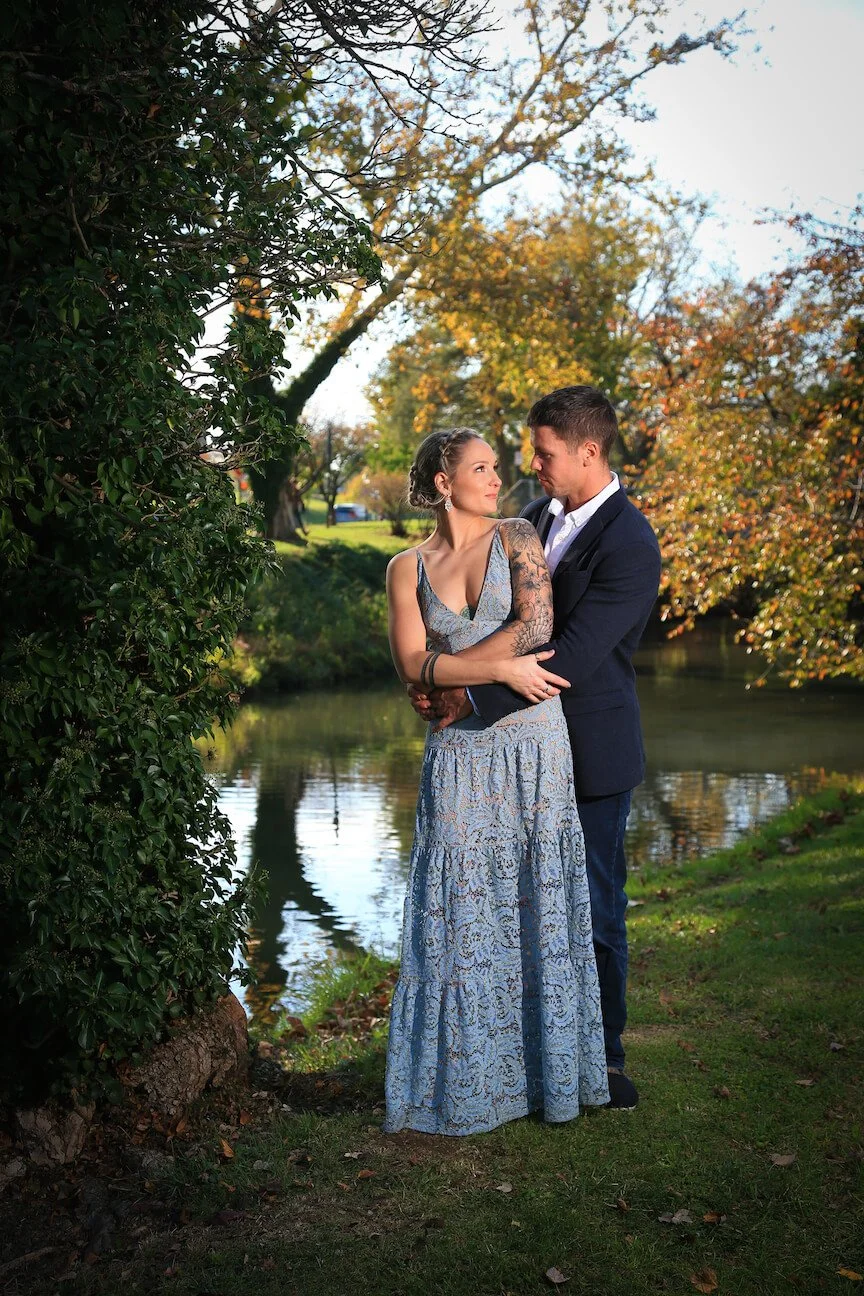 A couple standing close together by a pond in a park with autumn foliage, the woman wearing a long lace dress and the man in a dark suit, gazing into each other's eyes.