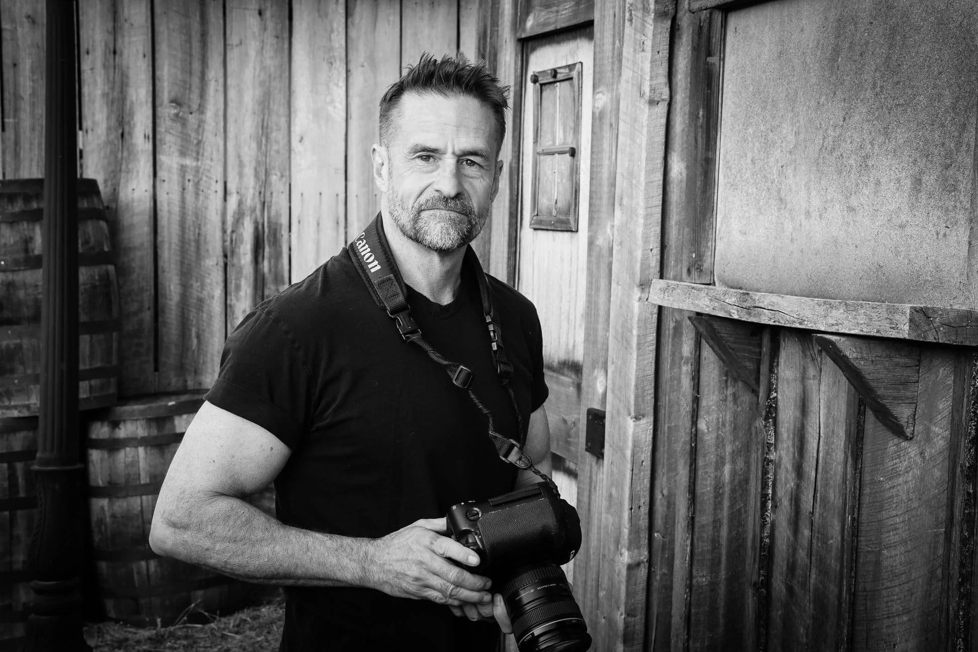 A man holding a camera stands in front of wooden walls, looking directly at the camera.