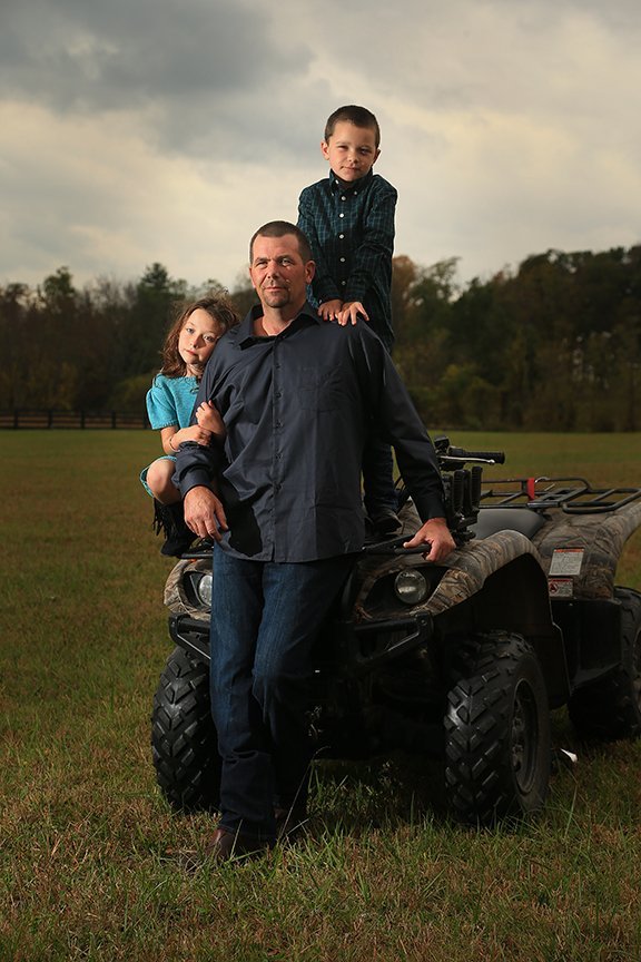 A man standing in front of an all-terrain vehicle with two children, a girl and a boy, sitting and standing on it outdoors in a grassy field with trees in the background.