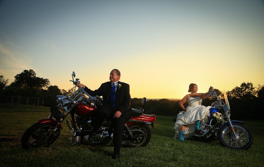 Bride and groom in wedding attire sitting on motorcycles in a field at sunset.