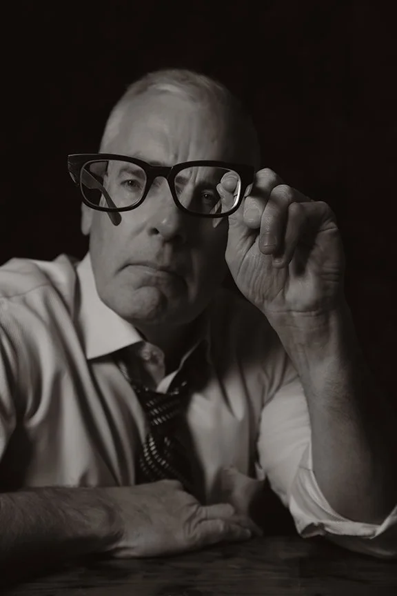 A black-and-white portrait of an elderly man wearing glasses, sitting at a table, adjusting his glasses with one hand and looking directly at the camera.
