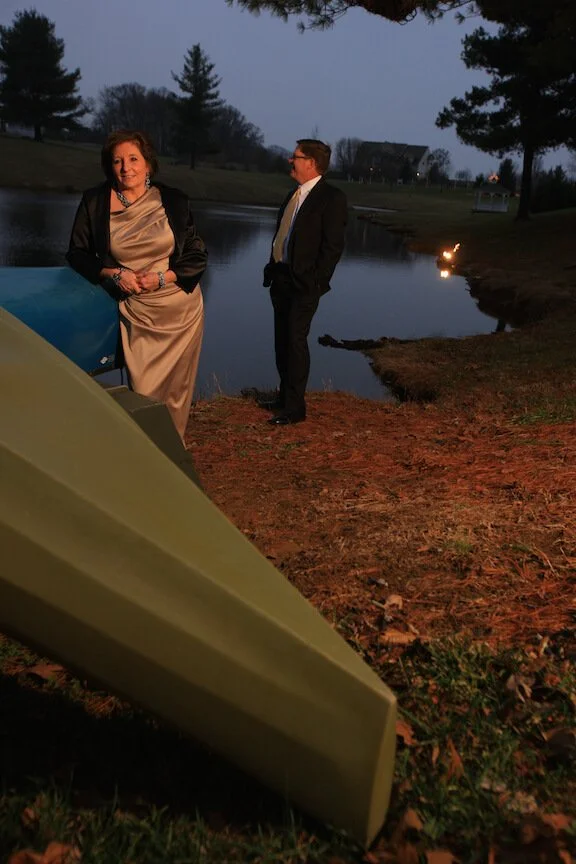 Two people dressed in formal attire standing by a pond during dusk, with a boat in the foreground and trees in the background.