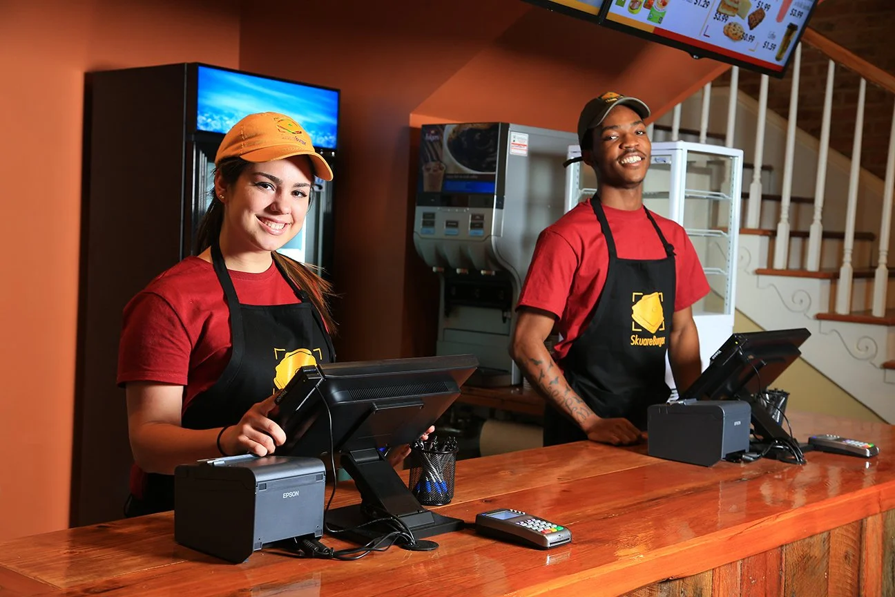 Two fast-food restaurant cashiers, a woman and a man, standing behind the counter wearing red shirts, black aprons with a yellow logo, and caps, smiling at the camera.