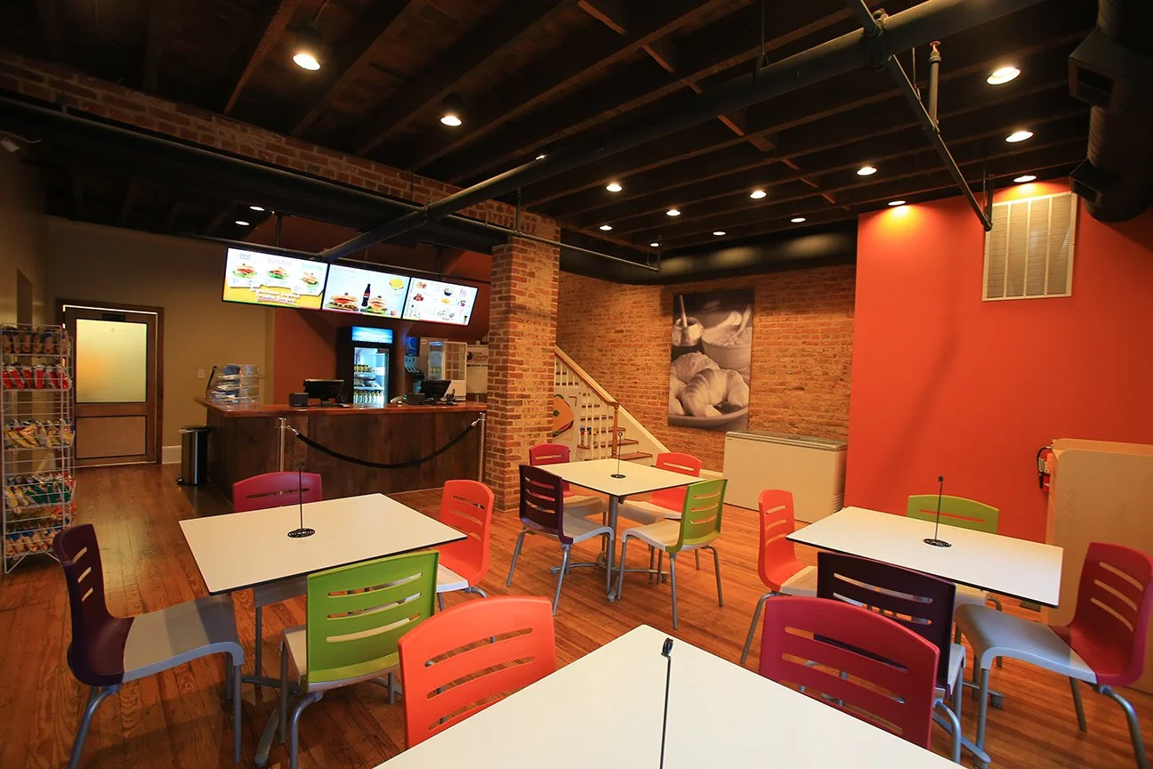 Empty colorful cafeteria with white tables, red, green, and purple chairs, a brick wall, a staircase, and a counter with digital menu screens.