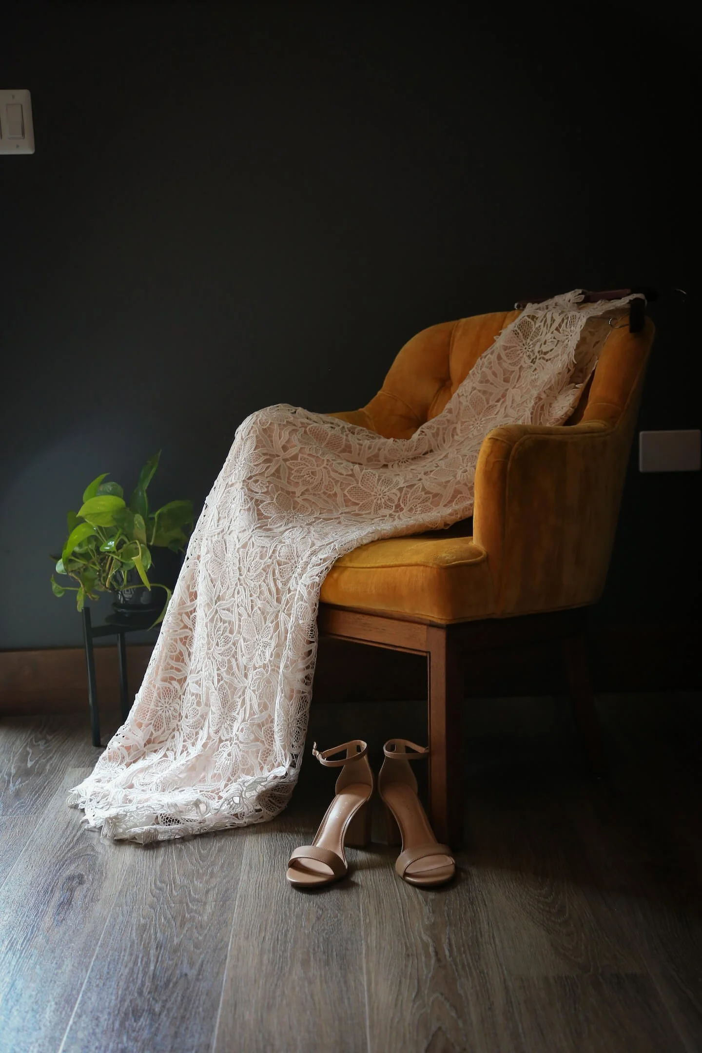 A vintage yellow armchair with a white lace dress draped over it, pair of beige high-heeled shoes on the floor in front, and a small green potted plant on a black stand next to it, set against a dark wall with wooden flooring.