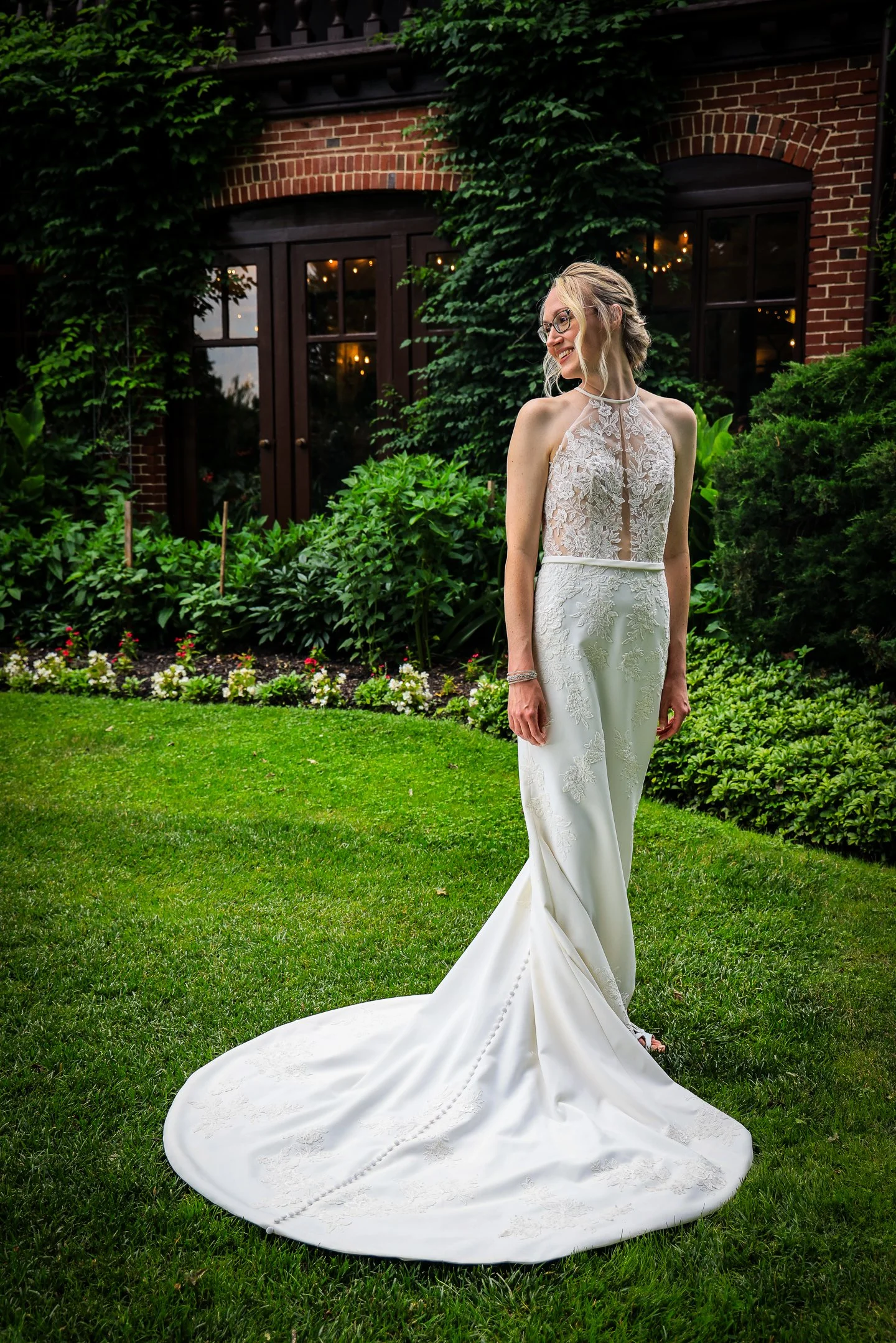 A bride in a lace wedding gown with a train standing in a lush garden with green shrubs and a brick building in the background.