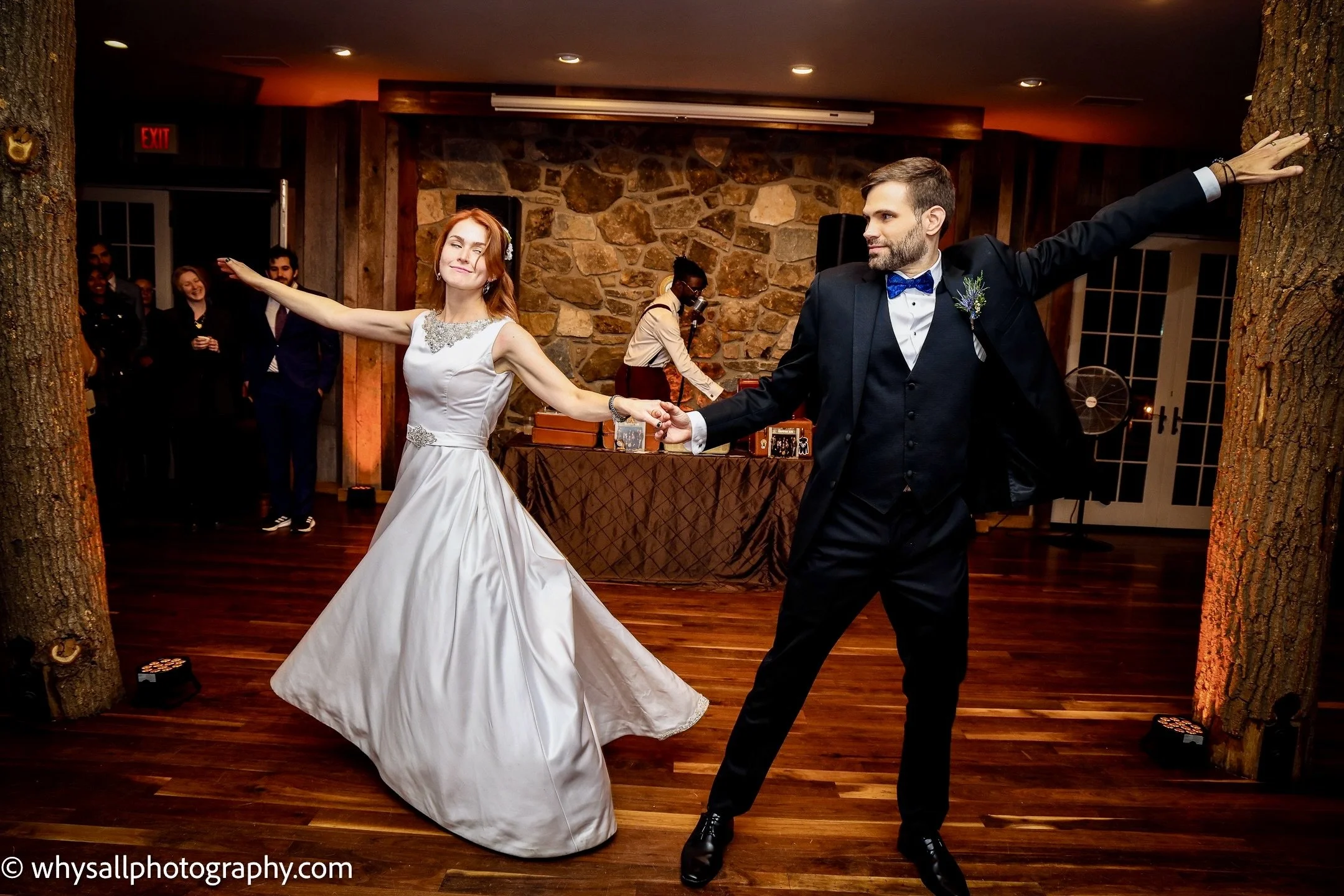 A bride and groom are dancing at their wedding reception, holding hands and smiling. The bride is wearing a white gown and the groom is in a tuxedo. There are guests in the background and a DJ or musician playing music behind them.