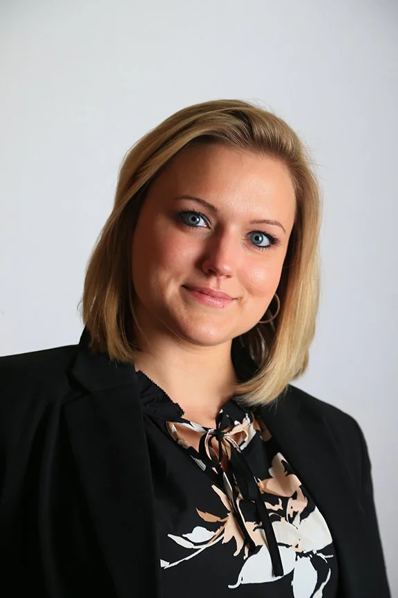 A young woman with blonde hair and blue eyes, smiling, wearing a black blazer and a patterned blouse with a bow, standing against a plain white background.