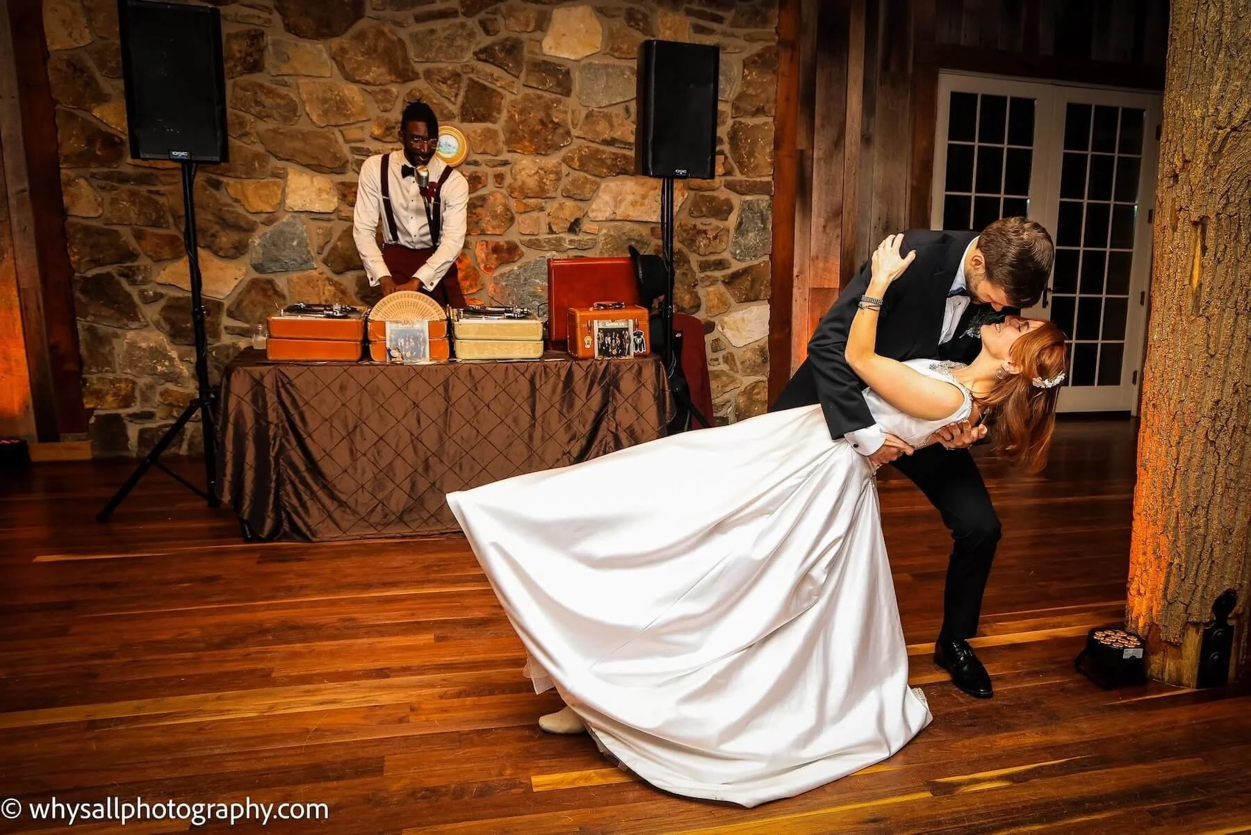 A bride and groom sharing a dance at their wedding reception, with a DJ playing music in the background and a stone wall.