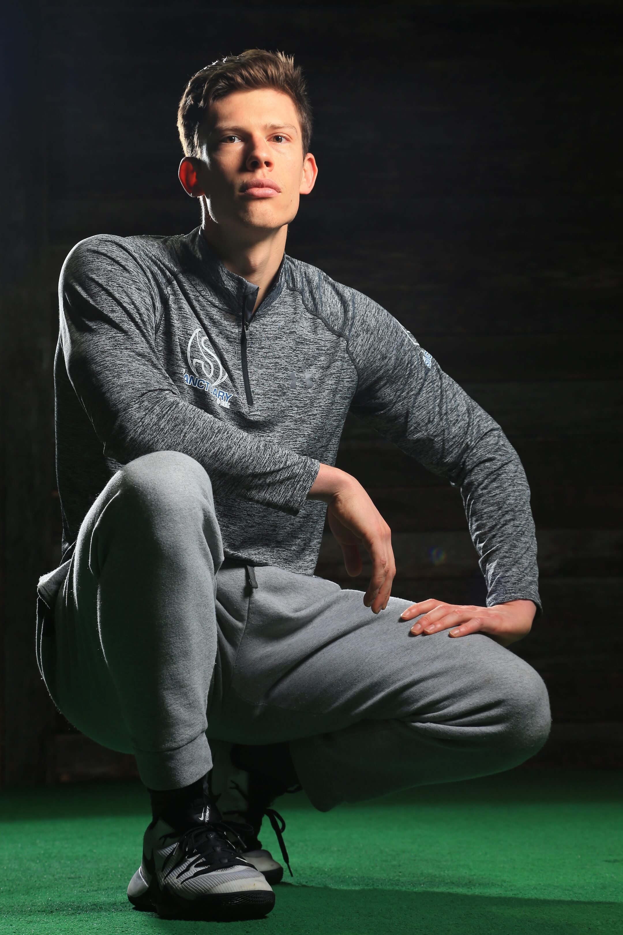 A young man in athletic clothing crouches on a green sports floor, with a dark background, looking confidently at the camera.