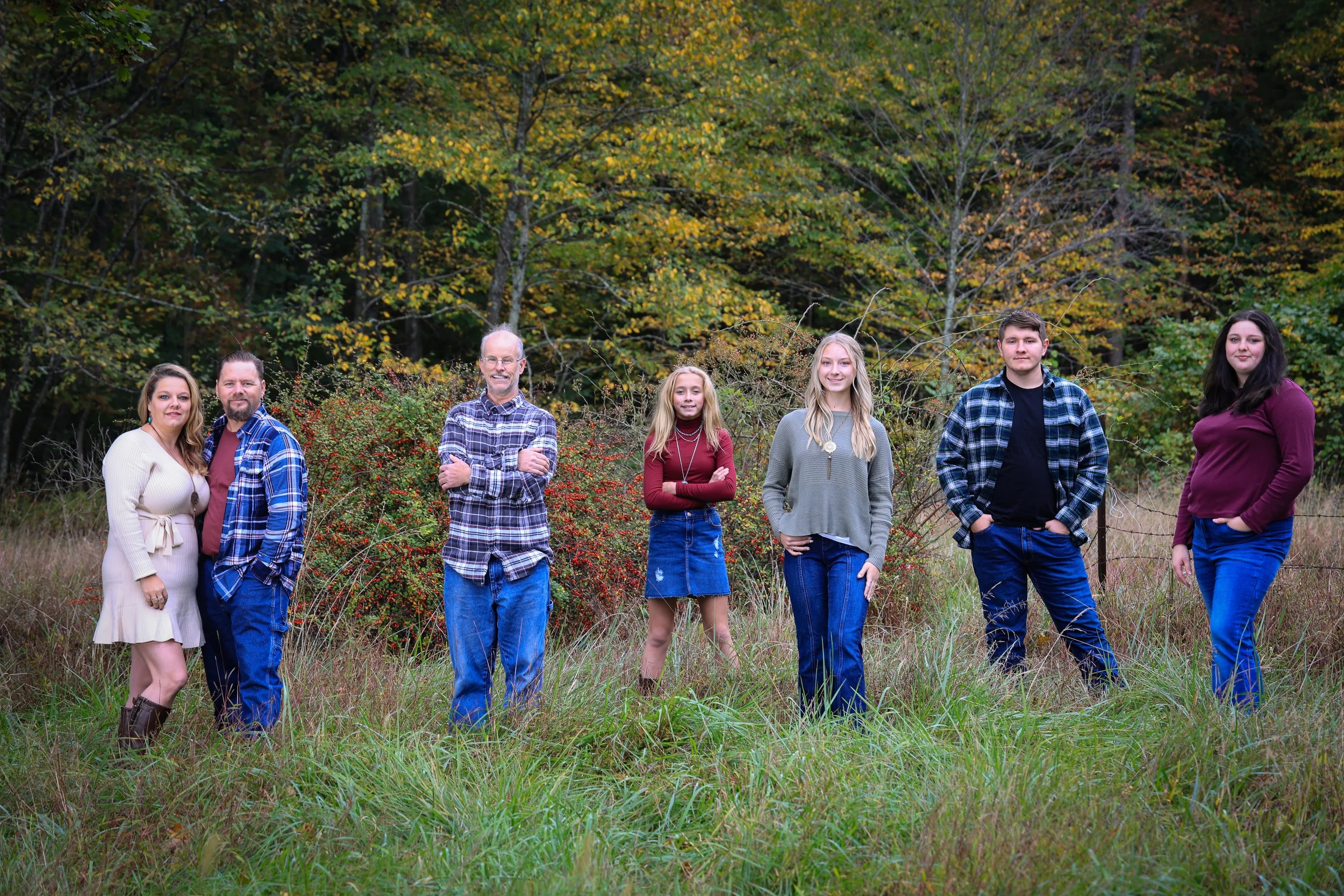 A family of seven standing outdoors in a grassy field with fall foliage trees in the background.