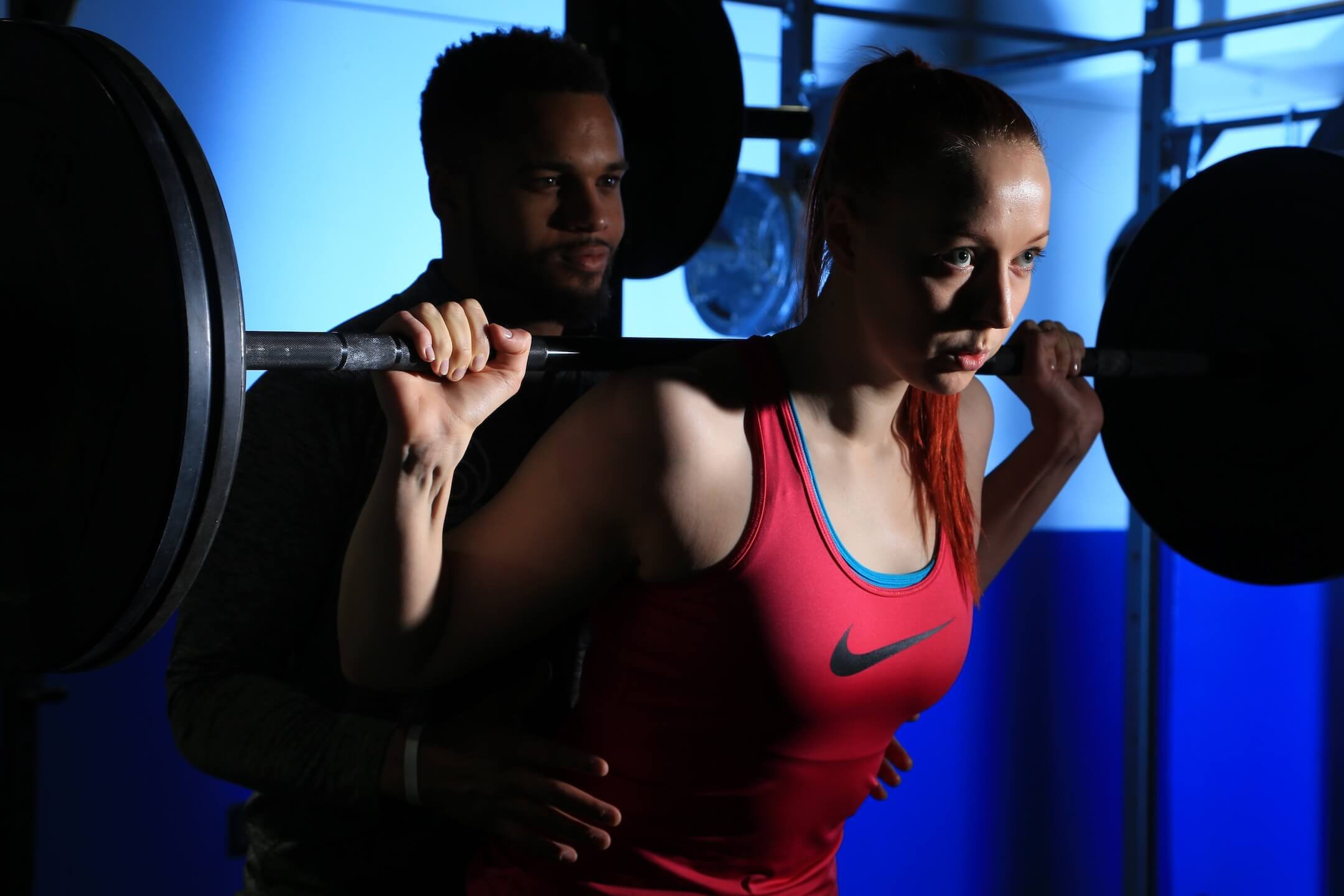 A woman in a pink sports top is performing a squat with a barbell on her shoulders, guided by a trainer behind her in a gym with blue lighting.