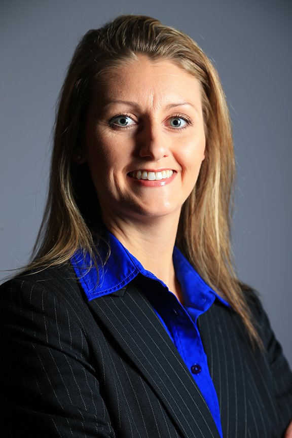 A professional woman with blonde hair, wearing a dark pinstripe suit and a bright blue shirt, smiling against a plain gray background.