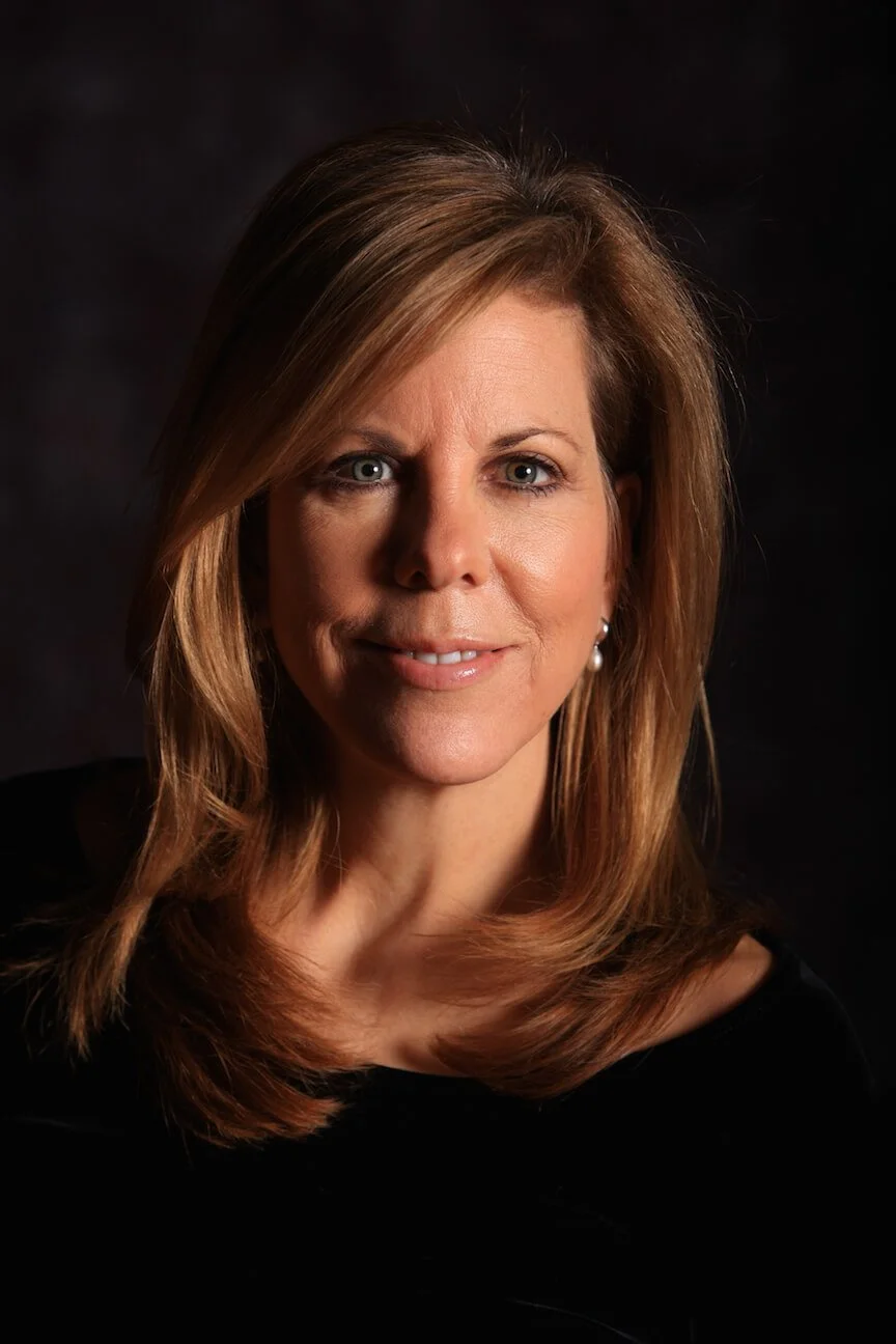 A woman with shoulder-length, light brown hair, blue eyes, and a slight smile, wearing a black top and a pearl earring, against a dark background.