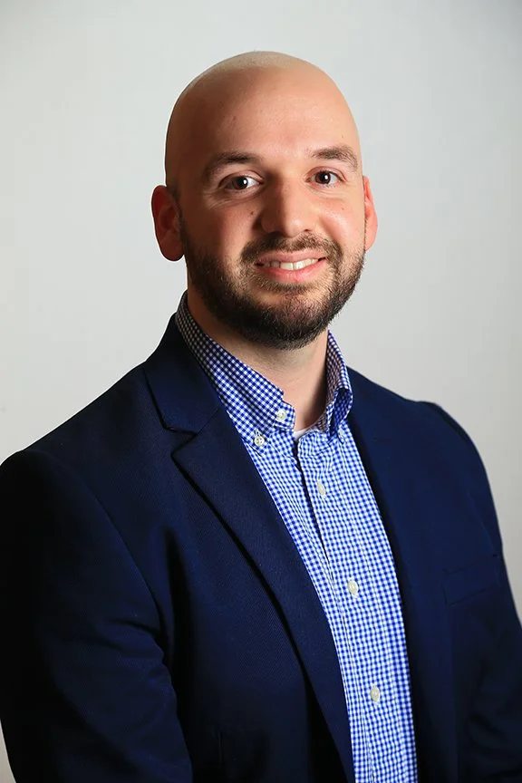 Headshot of a man with a bald head and beard, wearing a navy blazer and a blue checkered shirt, smiling against a plain background.
