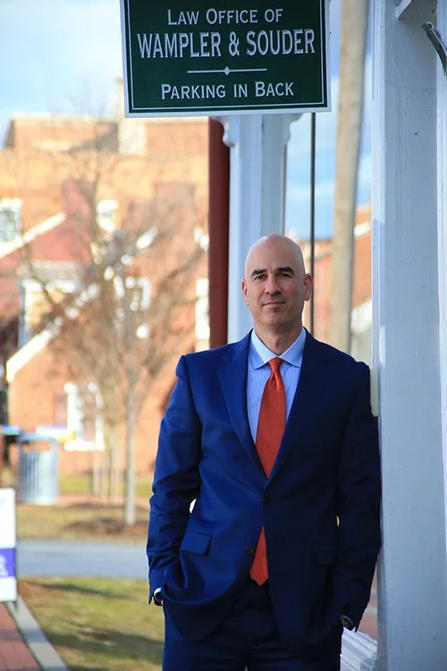 A bald man in a blue suit and orange tie stands outside a building with a sign that reads "Law Office of Wampler & Souder" and "Parking in Back." He has his hands in his pockets and is standing near a white wall, with a background of trees and buildi