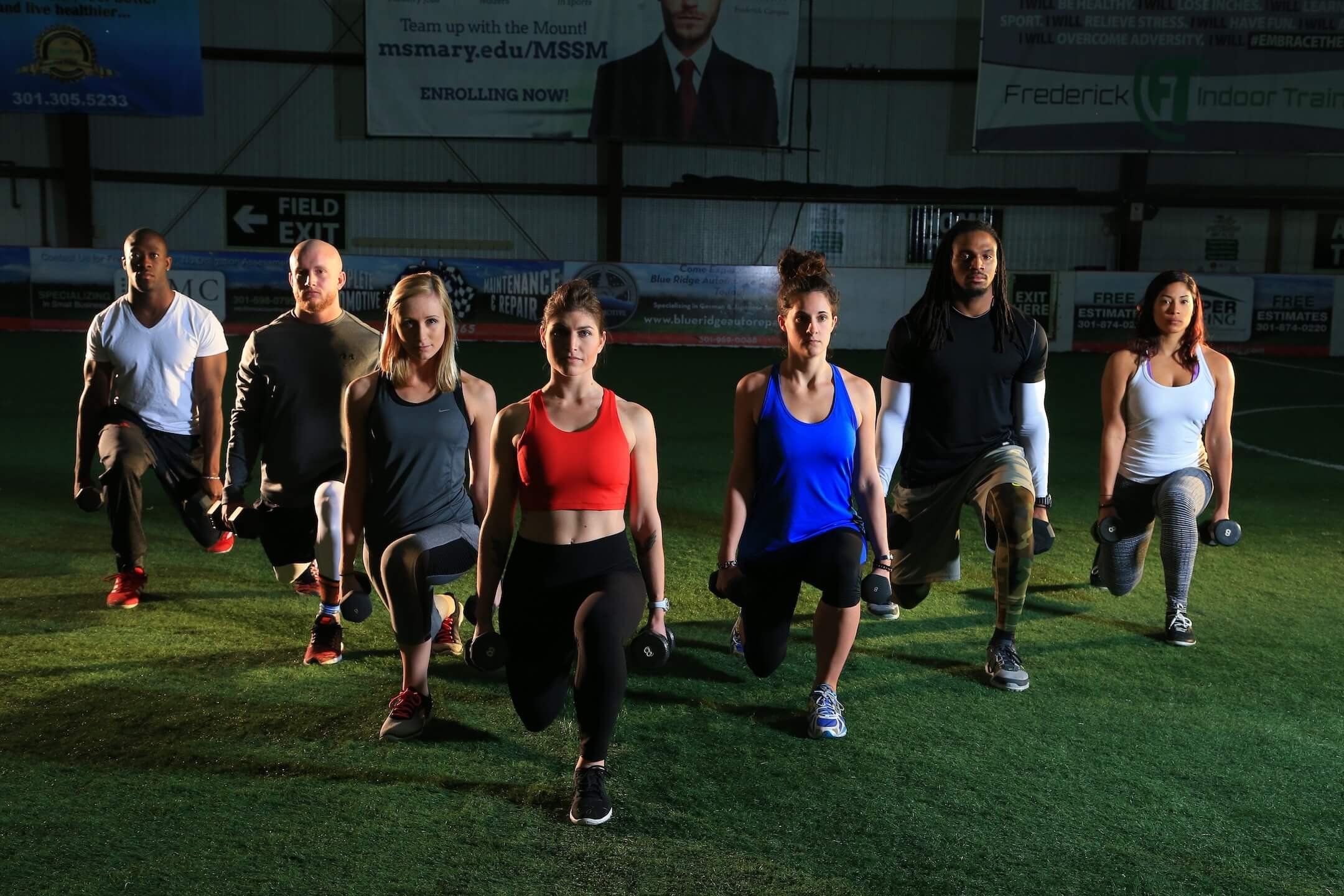 Group of seven diverse people exercising with dumbbells indoors on grass field.