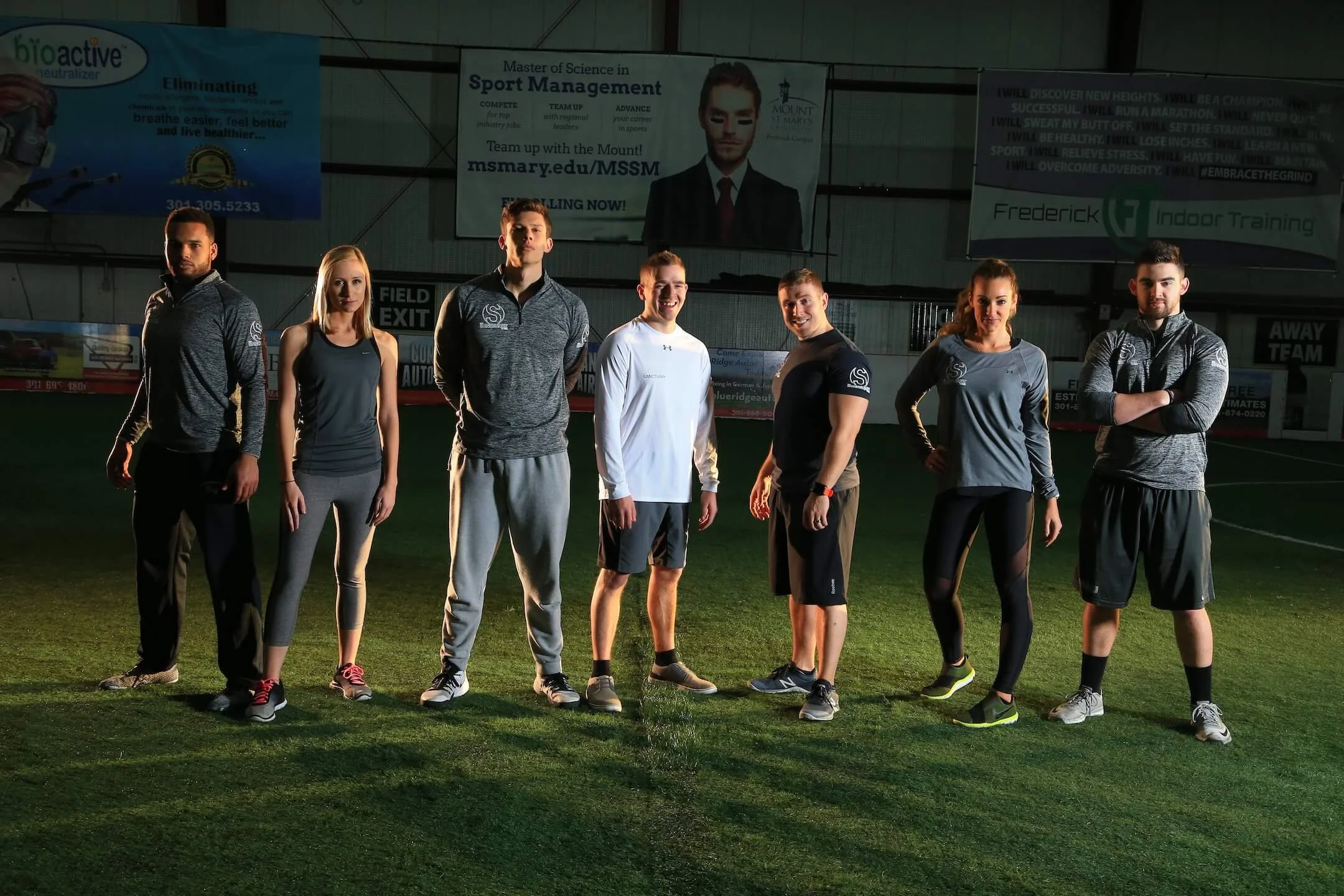 Group of seven athletes standing inside an indoor sports facility, with advertisements and banners on the wall behind them.