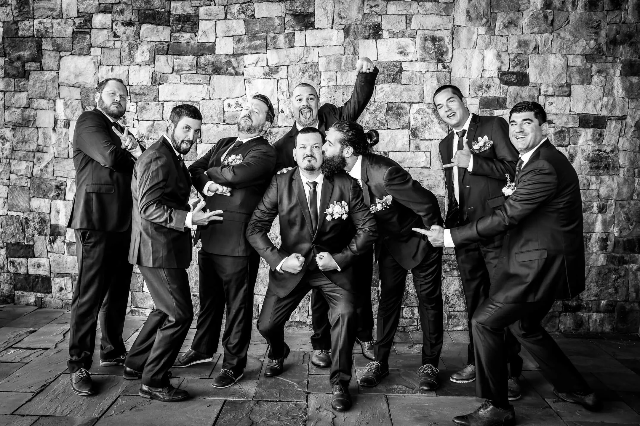 A group of nine men dressed in formal tuxedos posing together in front of a stone wall, with some making fun gestures and expressions.