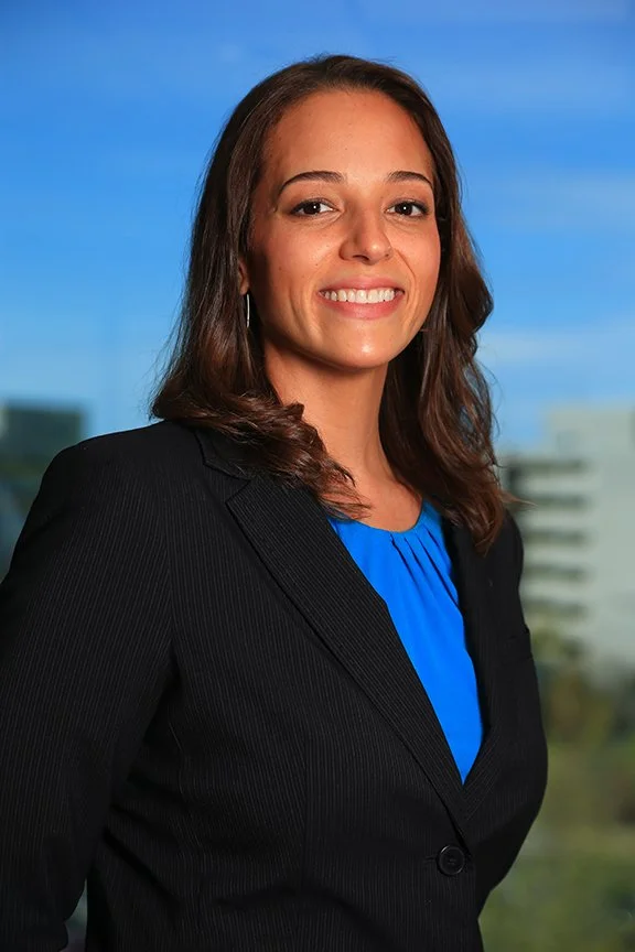 A woman in a black blazer over a blue blouse standing outdoors with a cityscape and blue sky in the background.