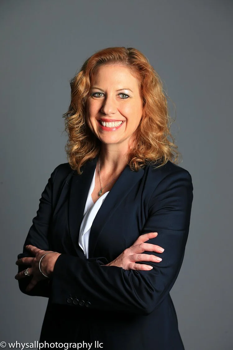 A woman with curly red hair wearing a dark blazer and white blouse, smiling with arms crossed against a plain gray background.