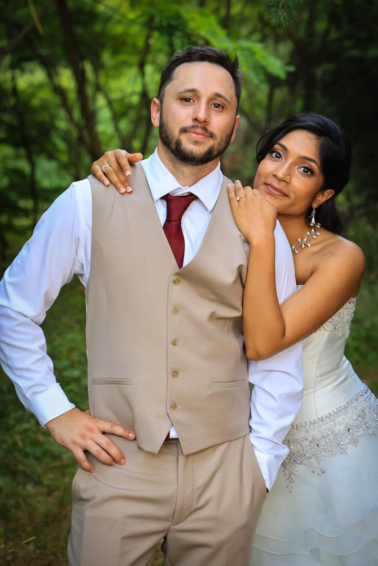 A couple in wedding attire posing outdoors with lush greenery in the background.