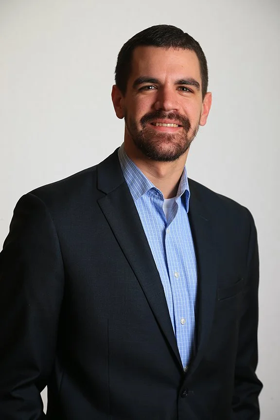 A man with short dark hair and a beard, wearing a dark suit jacket and a light blue dress shirt, smiling at the camera against a plain white background.