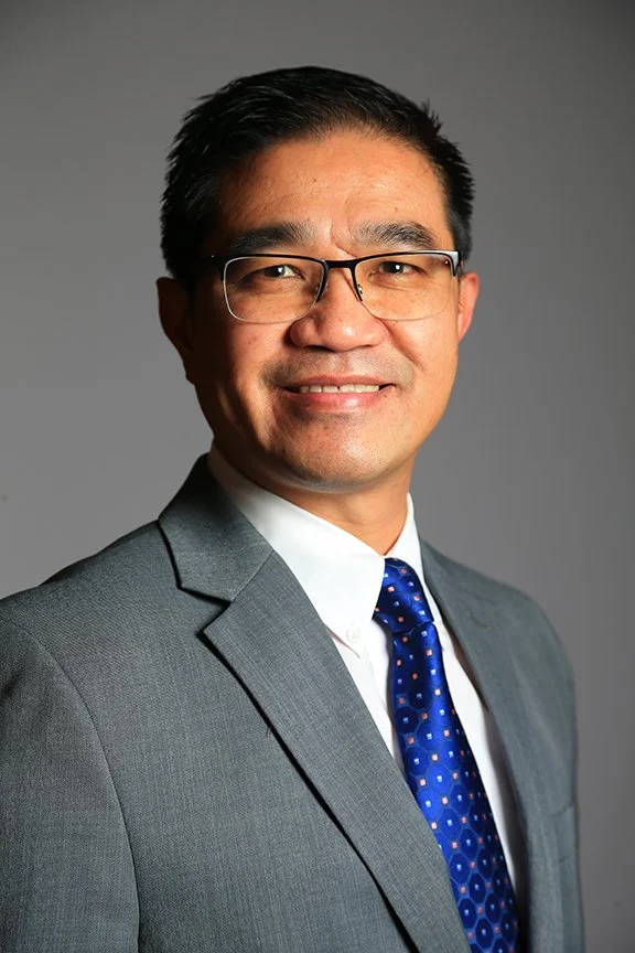 Professional portrait of an Asian man wearing glasses, a gray suit, white shirt, and a blue tie with small patterns, smiling against a neutral gray background.
