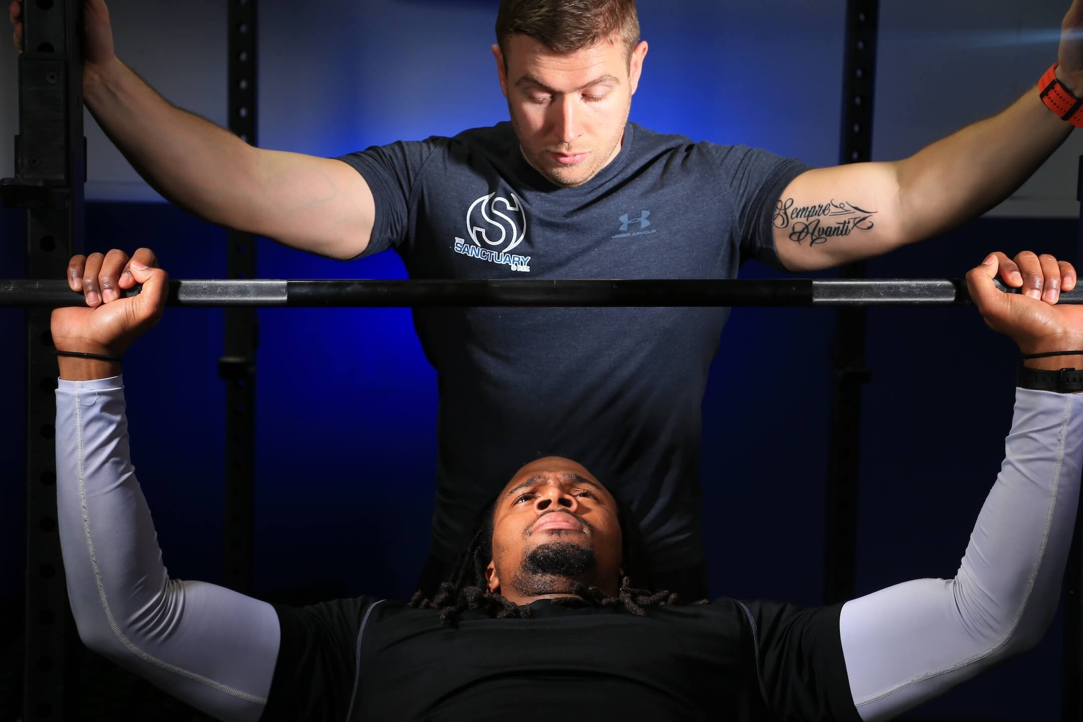 Two men are in a gym; one is lying on a bench pressing a barbell, and the other is standing above him spotting him, both focused on the exercise.