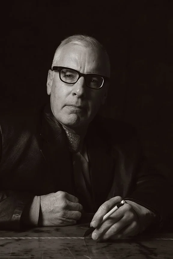 A black and white photo of a man with glasses sitting at a table, holding a pen, wearing a suit and tie, with a serious expression.