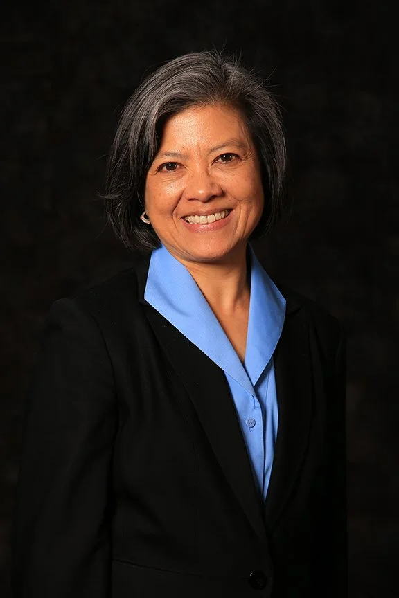 Smiling woman with short gray hair, wearing a black blazer and light blue shirt against a dark background.