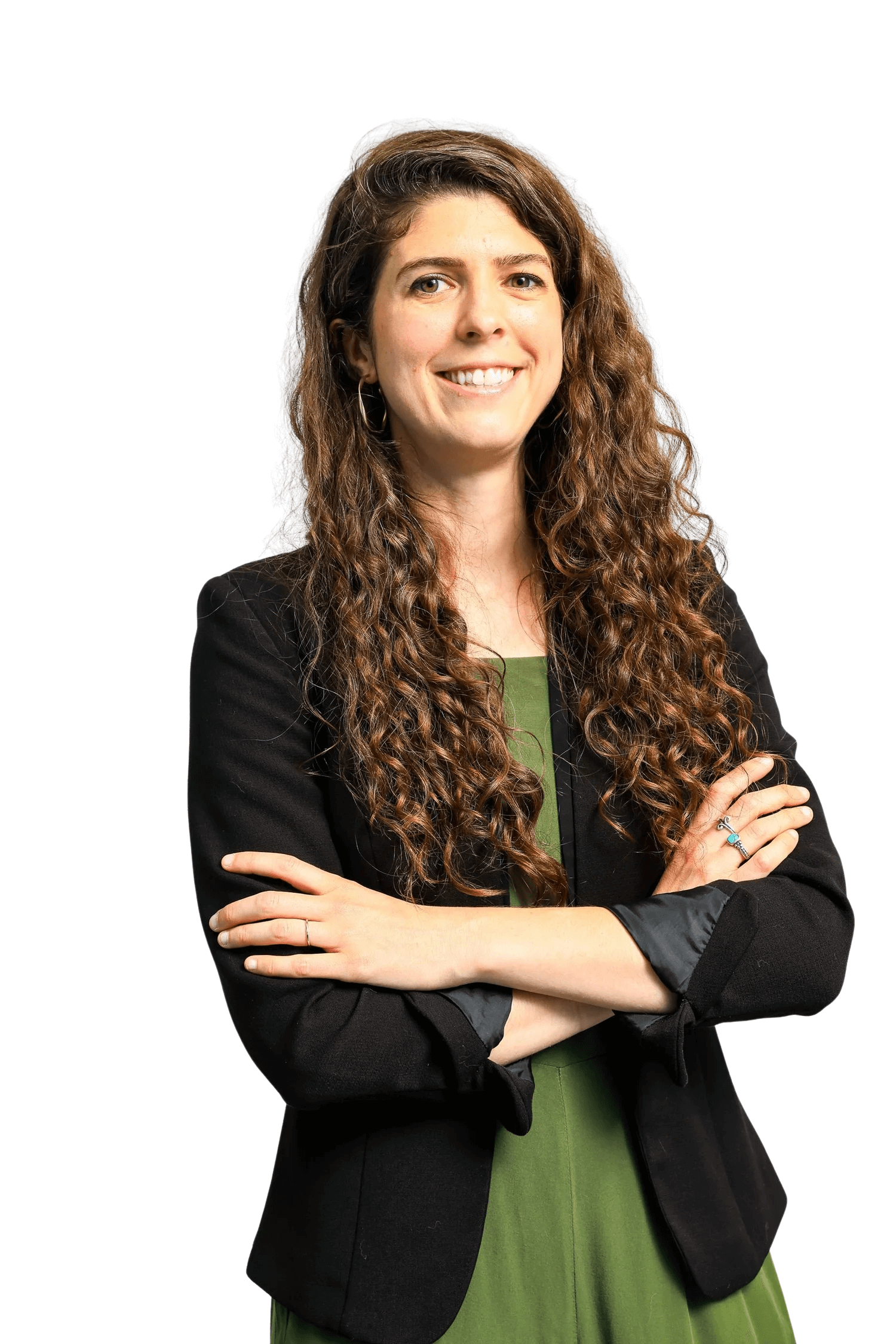 A woman with long curly brown hair, wearing a black blazer over a green dress, standing with arms crossed and smiling at the camera against a white background.