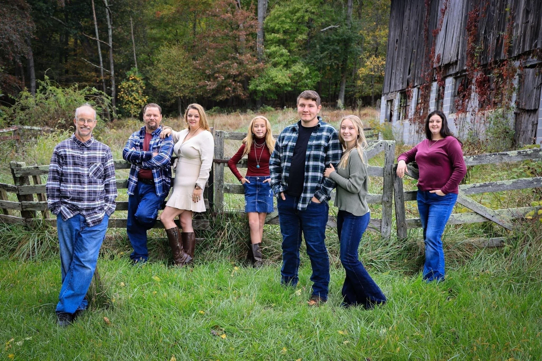 Family of seven standing outdoors near a wooden fence and old barn with fall foliage in the background.