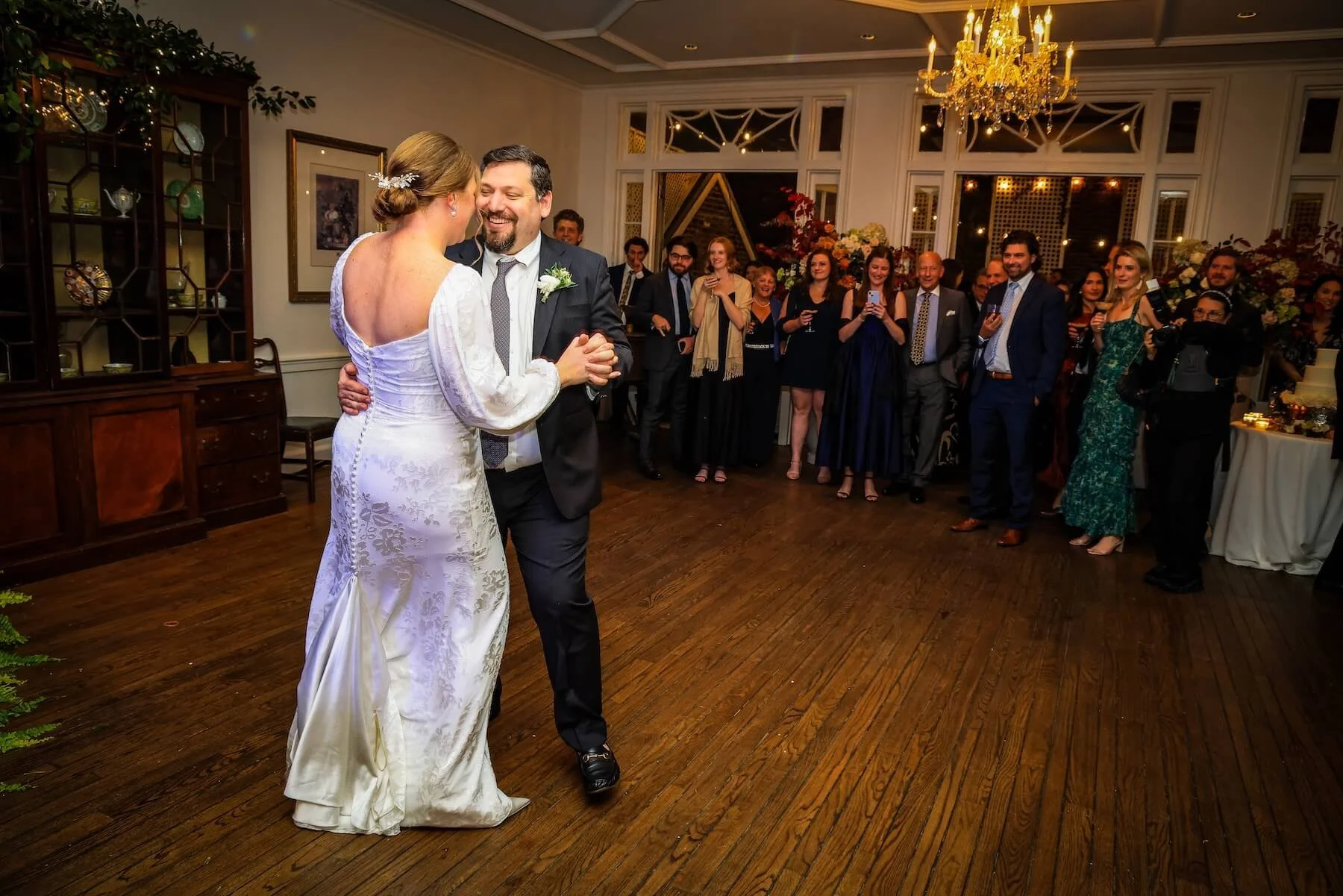 A bride and groom share a dance in the center of a wedding reception, with guests forming a semi-circle around them, smiling and capturing the moment.