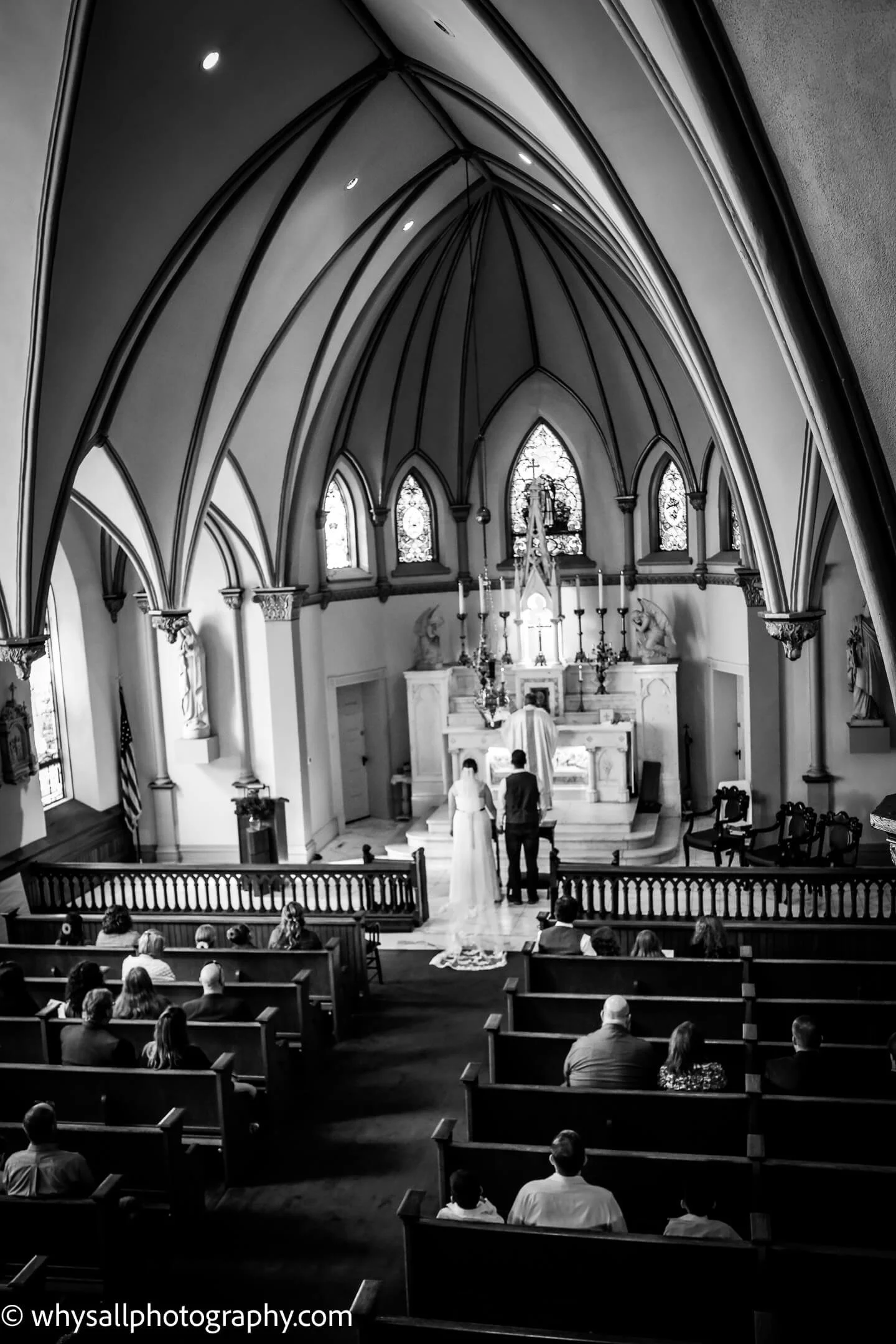 A wedding ceremony inside a church, with a bride and groom standing at the altar, under vaulted ceilings with Gothic arches, and guests seated in pews watching.