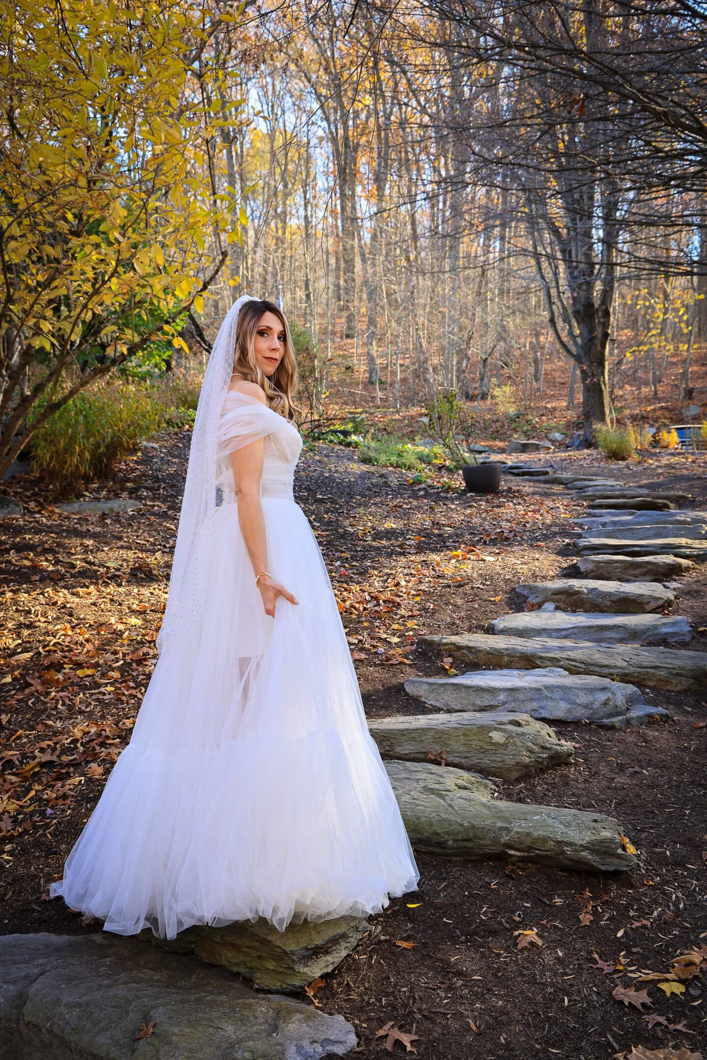 A woman dressed in a white wedding gown with a veil stands outdoors on a stone path in a wooded area during autumn, with trees showing fall foliage in the background.