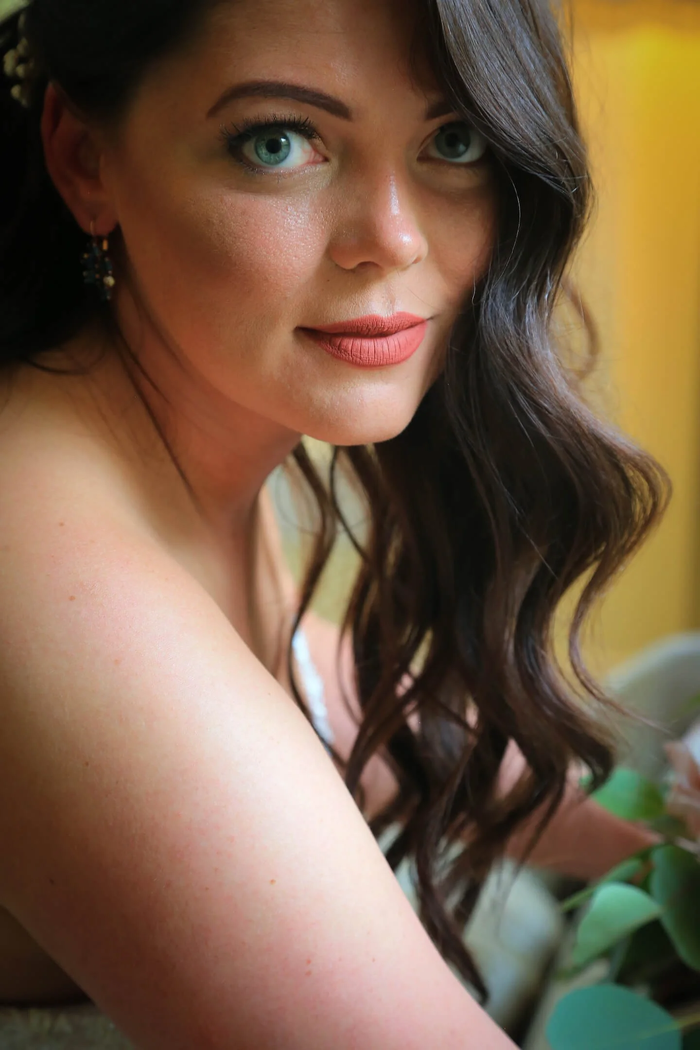 Close-up of a woman with blue eyes and dark brown wavy hair, wearing earrings and makeup, with a soft yellow background.