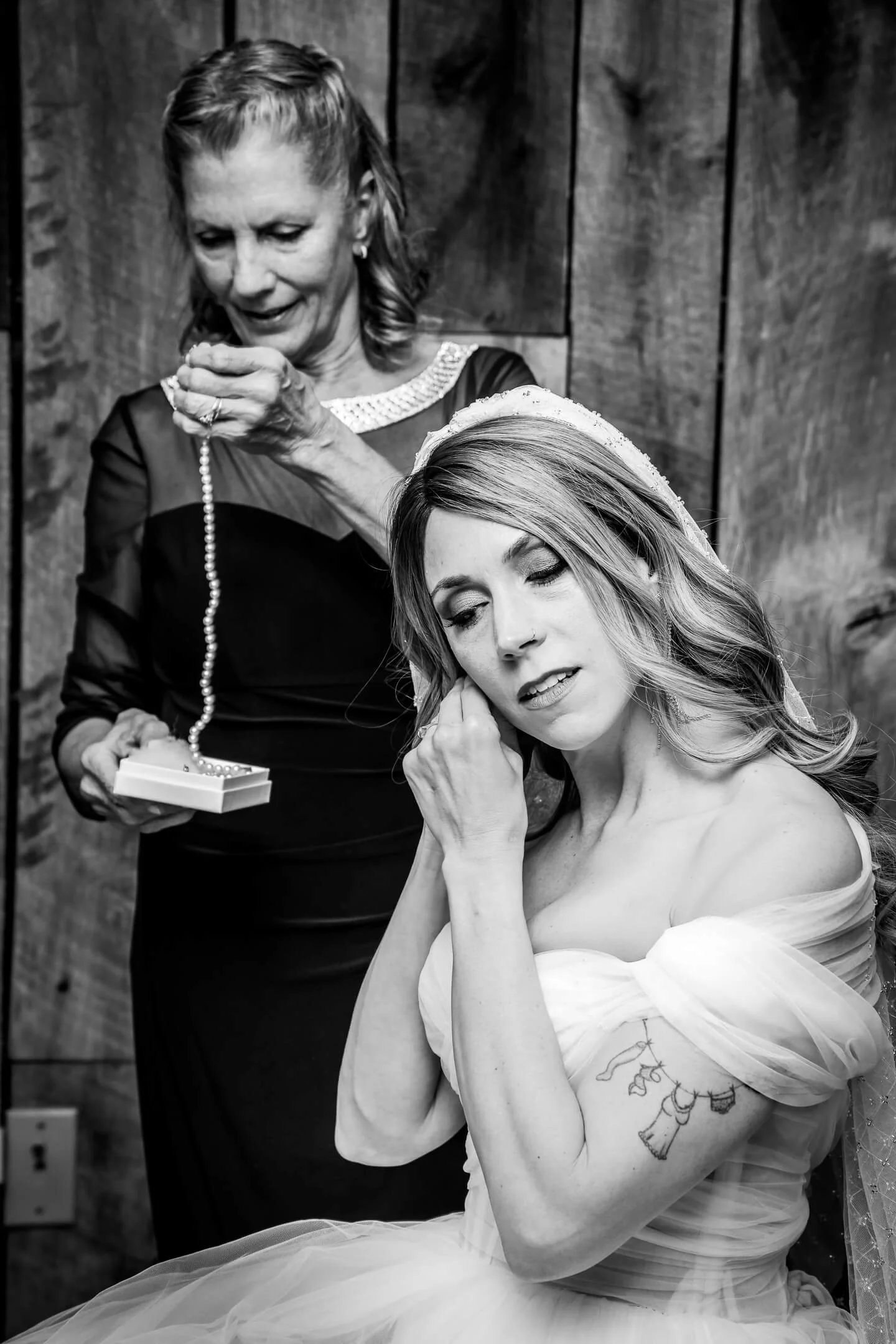 A bride with long, wavy hair and a tattoo on her right arm, sits with her eyes closed and hand near her ear, tradition of the bridal veil, as an older woman in a dark dress holds a jewelry box and looks down at a necklace, the backdrop is wooden paneling.