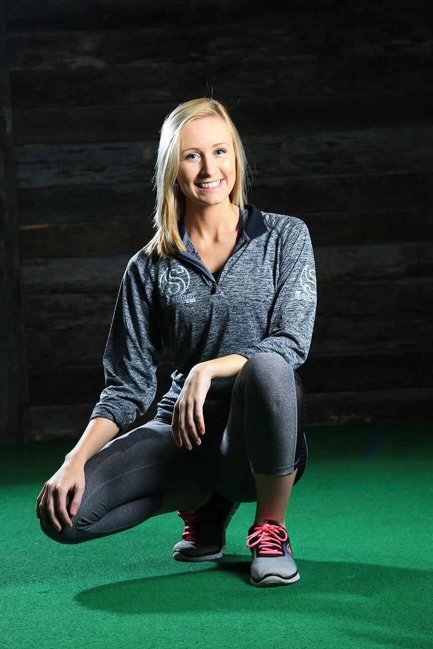 A smiling woman with blonde hair wearing a gray athletic jacket and black leggings, kneeling on one knee on a green gym floor with a dark wooden wall in the background.