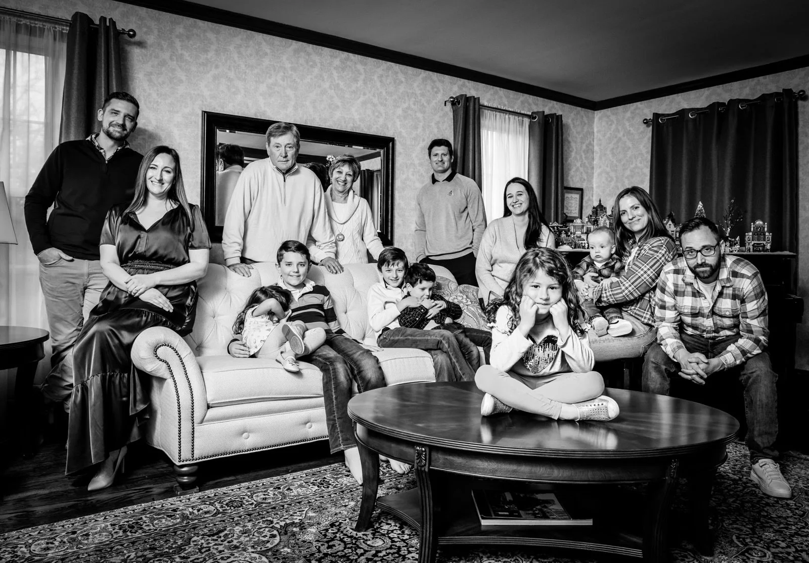 A family photo of fifteen people, including children and adults, gathered in a living room with patterned wallpaper, curtains, and a decorated background. They are posing and smiling.
