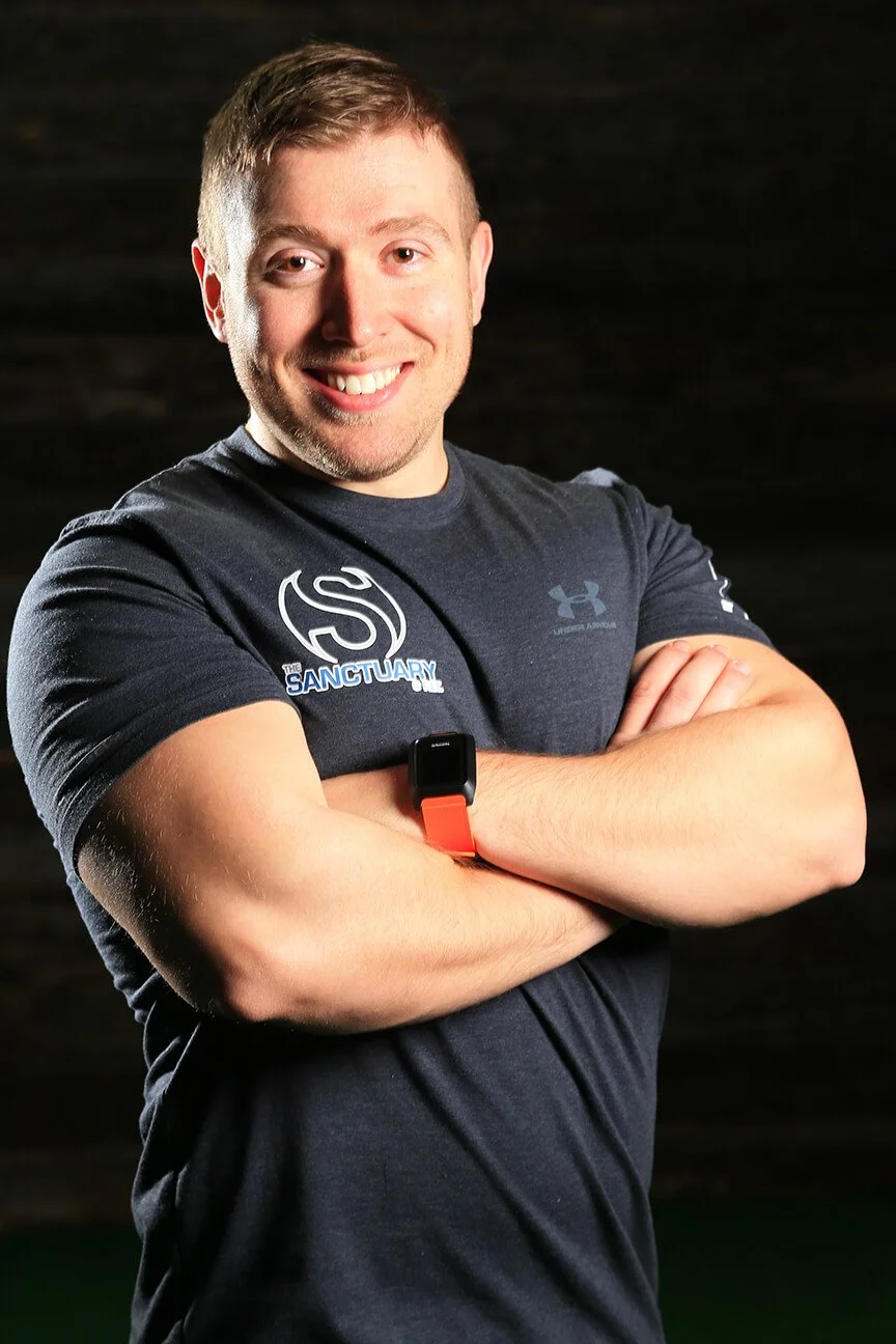 A young man with short light brown hair, smiling and crossing his arms, wearing a dark gray Under Armour t-shirt with logos, an orange fitness tracker on his left wrist, against a dark background.