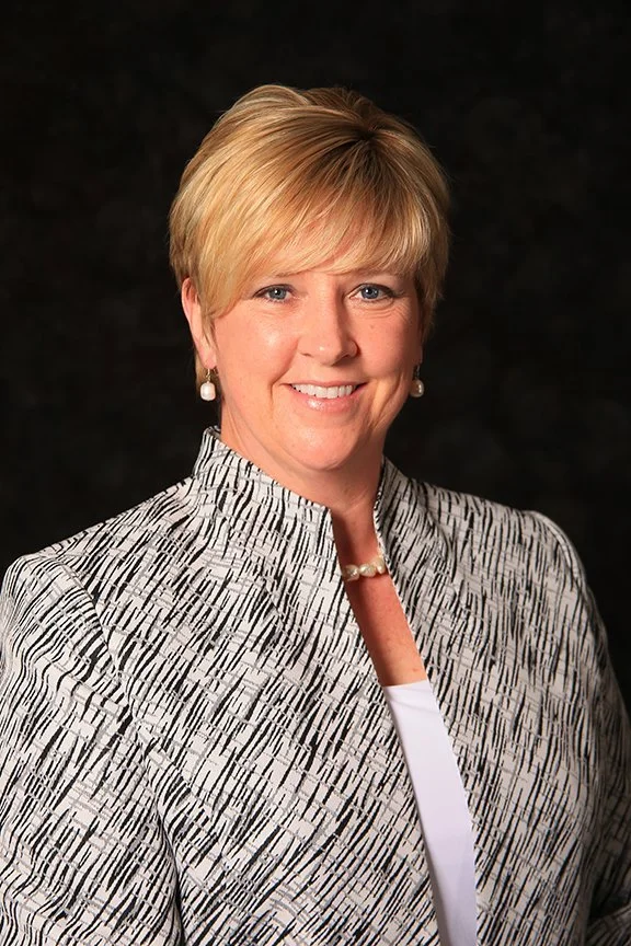 A professional woman with short blonde hair, wearing pearl earrings, a white top, and a patterned blazer, smiling against a dark background.